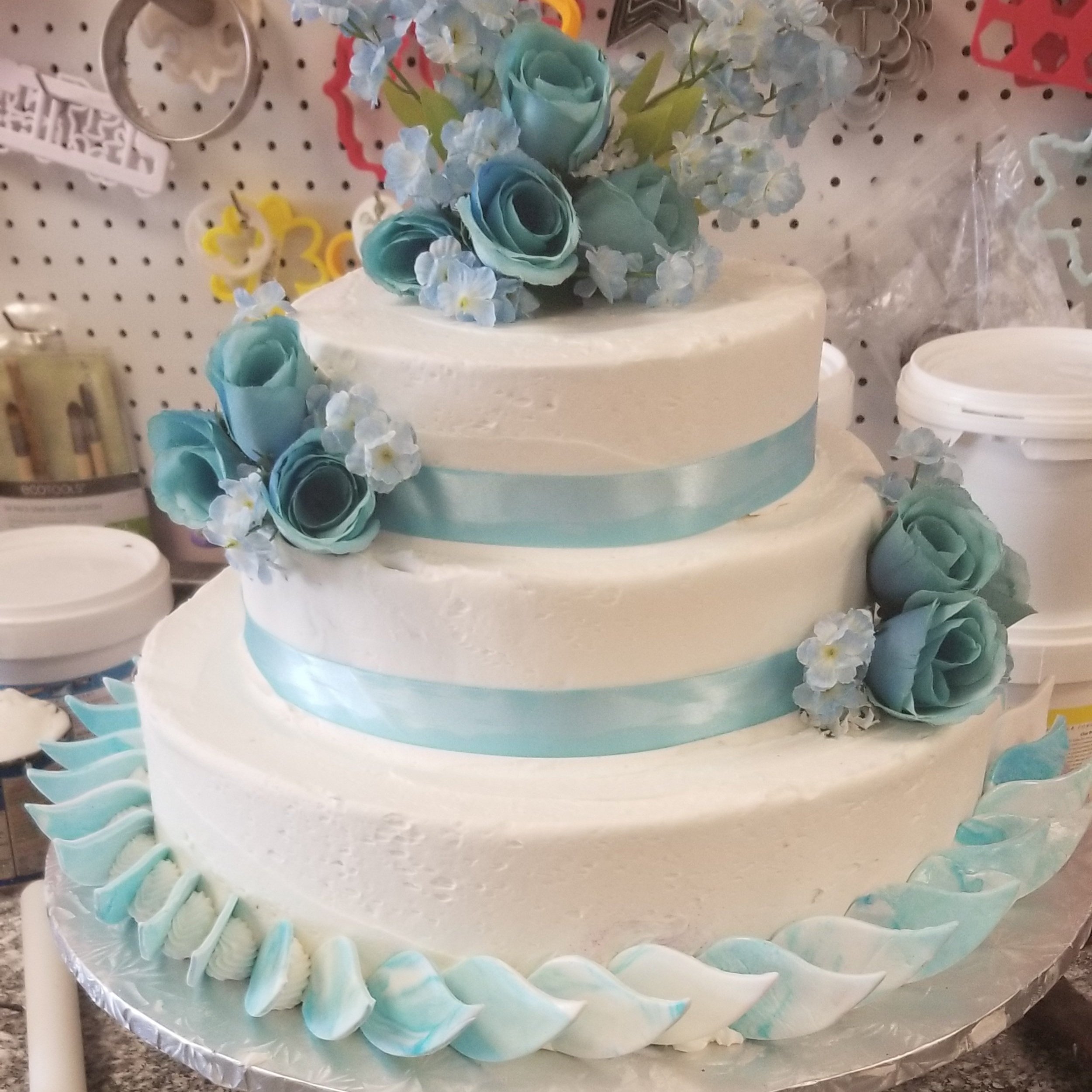 A three-tiered white wedding cake decorated with light blue ribbons and topped with light blue roses and small white flowers, sitting on a silver-colored base.