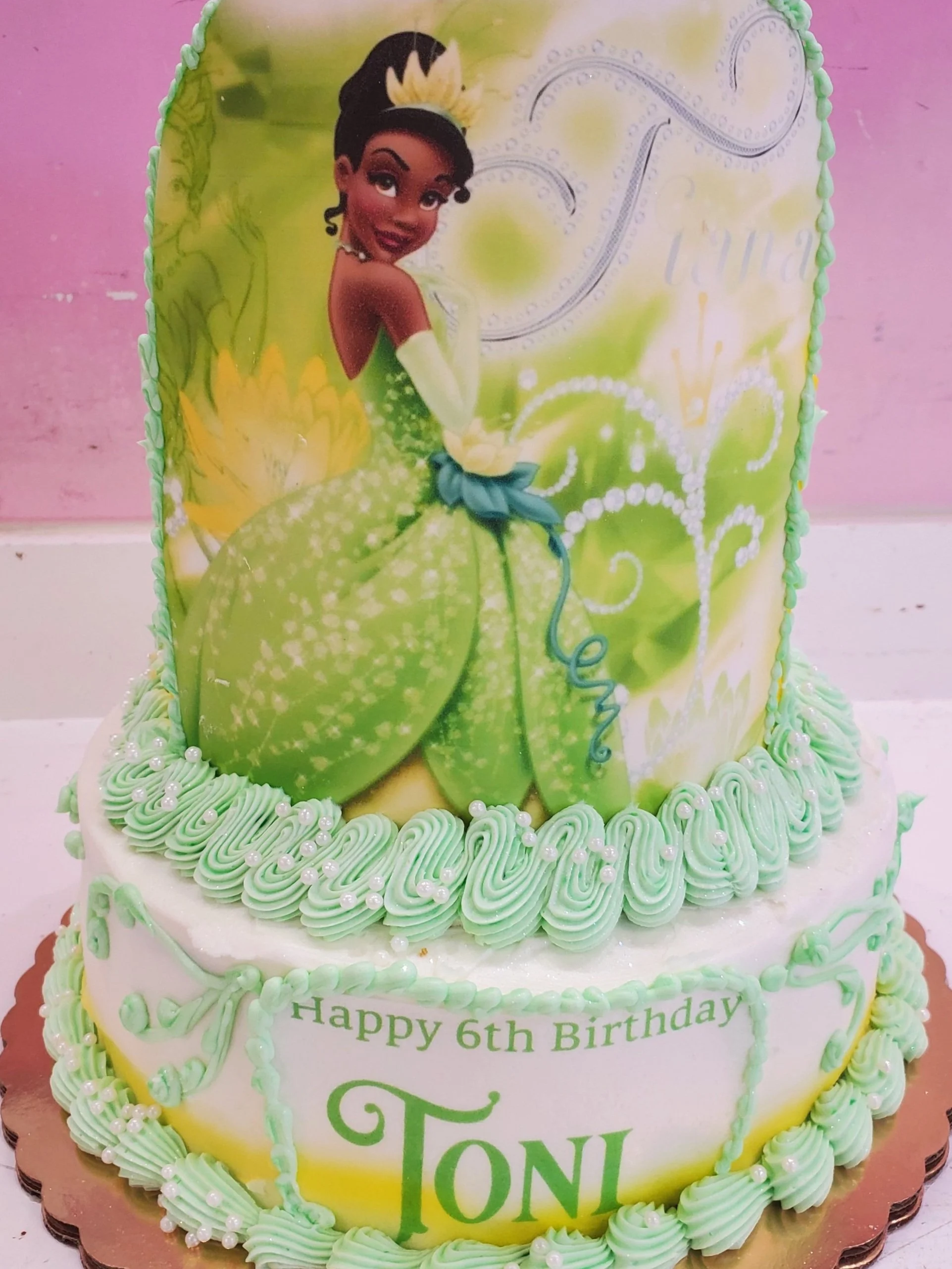 A two-tier birthday cake featuring a Tiana design. The top tier has an edible image of Tiana in a green gown, and the bottom tier is decorated with green and white icing with text that reads "Happy 6th Birthday TONI". The cake is decorated with piped icing and small pearl-like accents.