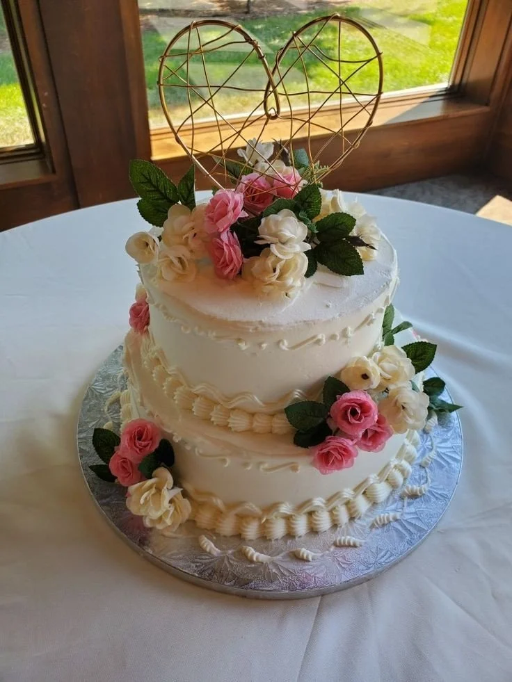 Two-tier white wedding cake decorated with pink and white flowers and a gold wire heart topper. 