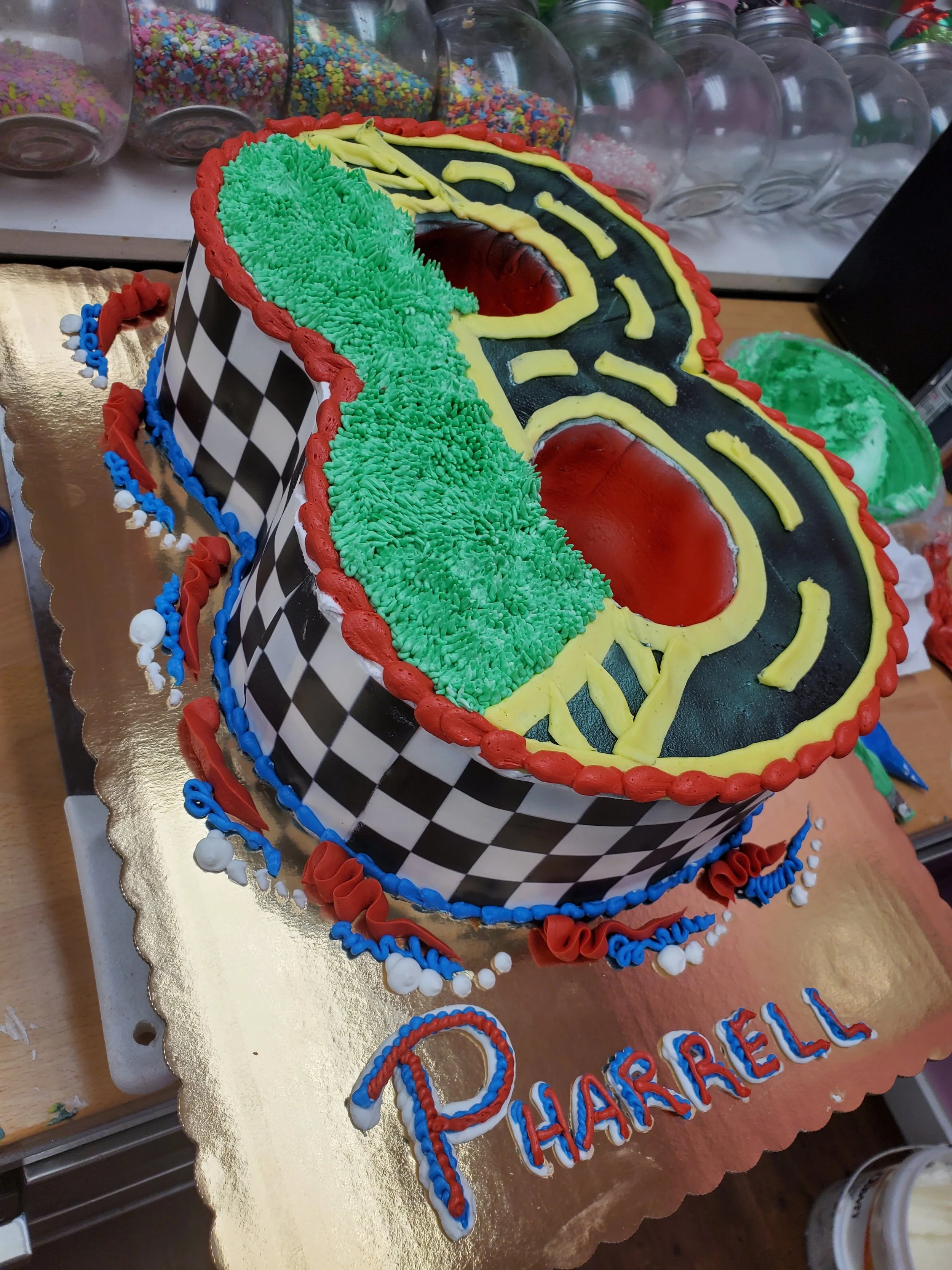 Race car themed birthday cake with colorful frosting, checkered pattern, and the name 'Pharrell' written on the cake board.