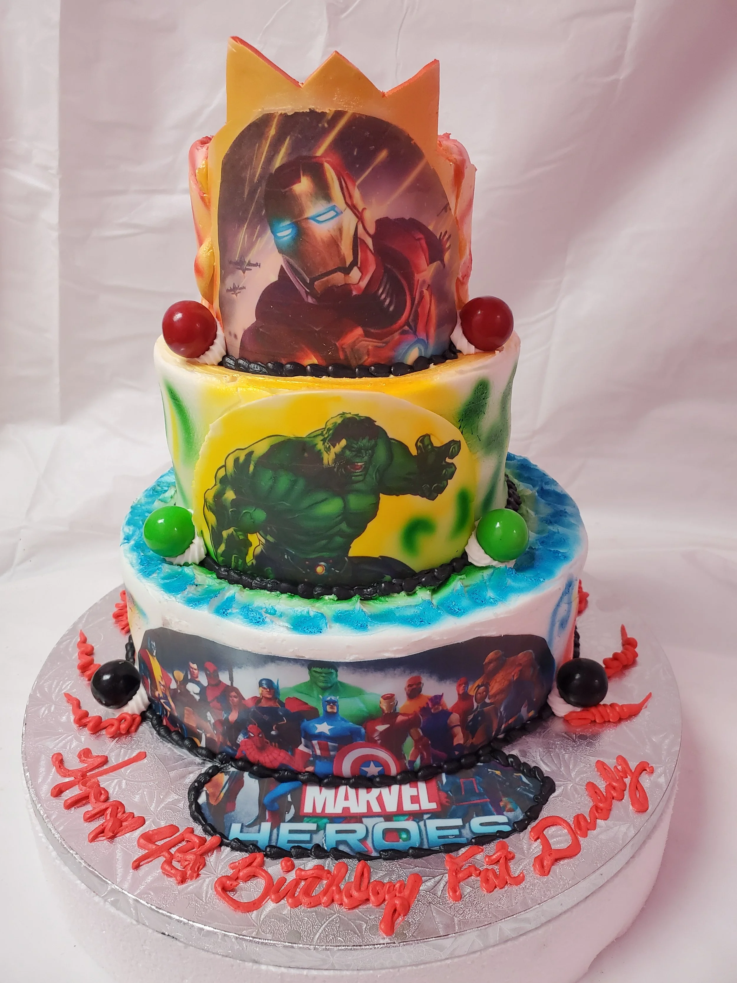 Three-tier Marvel Avengers themed birthday cake with superhero images, colorful icing, and red, green, black, and red decorative balls on a silver cake board. The message in red icing reads "Happy 4th Birthday Fat Dadddy."