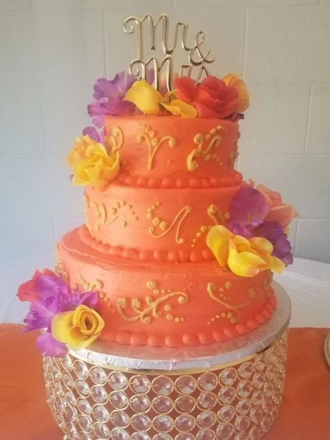 A three-tier pink and peach wedding cake decorated with yellow, purple, and orange flowers. The cake features Lambeth Piping. A WILD One!