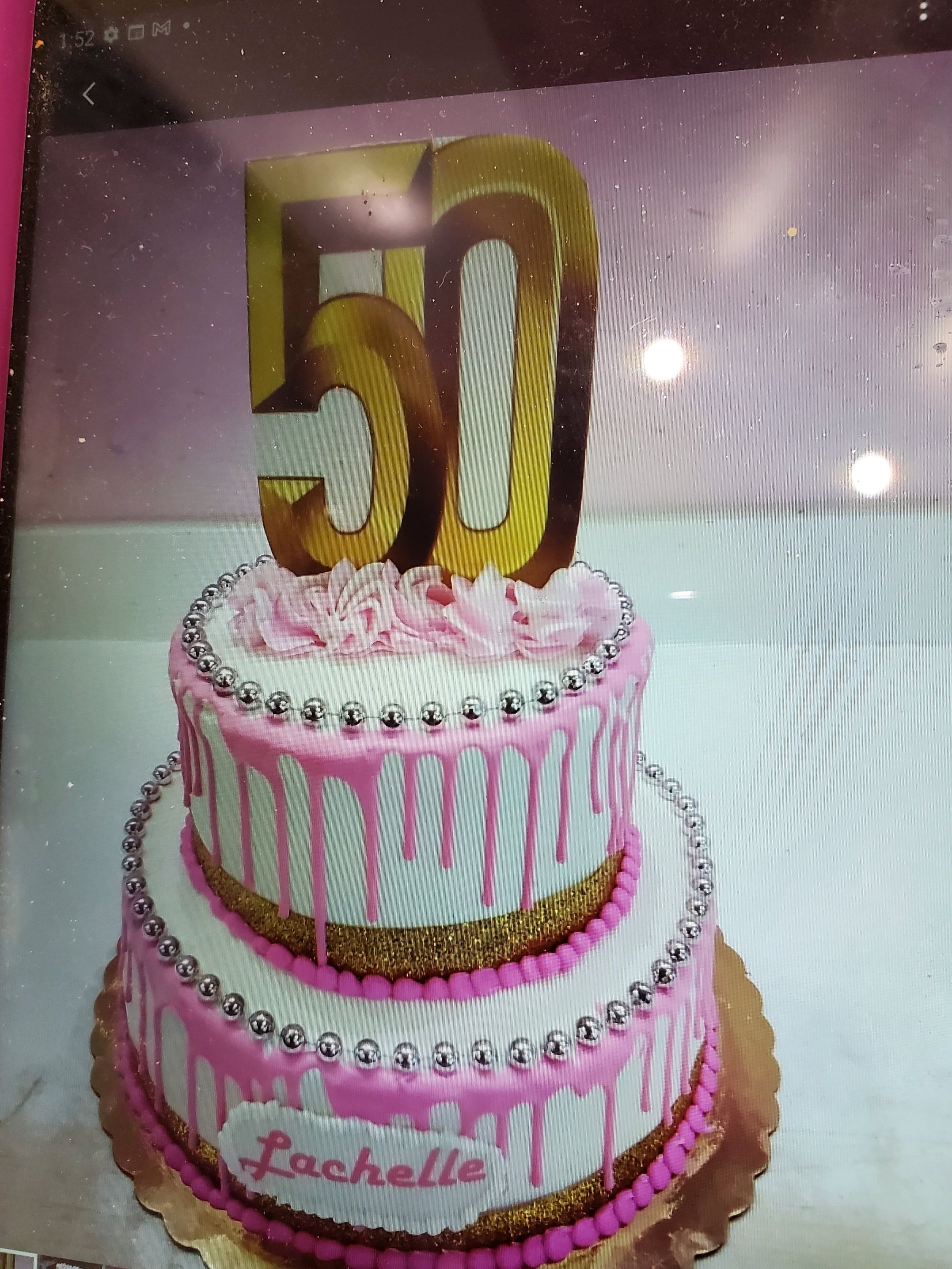 Pink and white birthday cake with a large gold '50' topper, decorated with pink icing drips, silver beads, and gold accents, with a heart-shaped plaque that says "Lachelle."