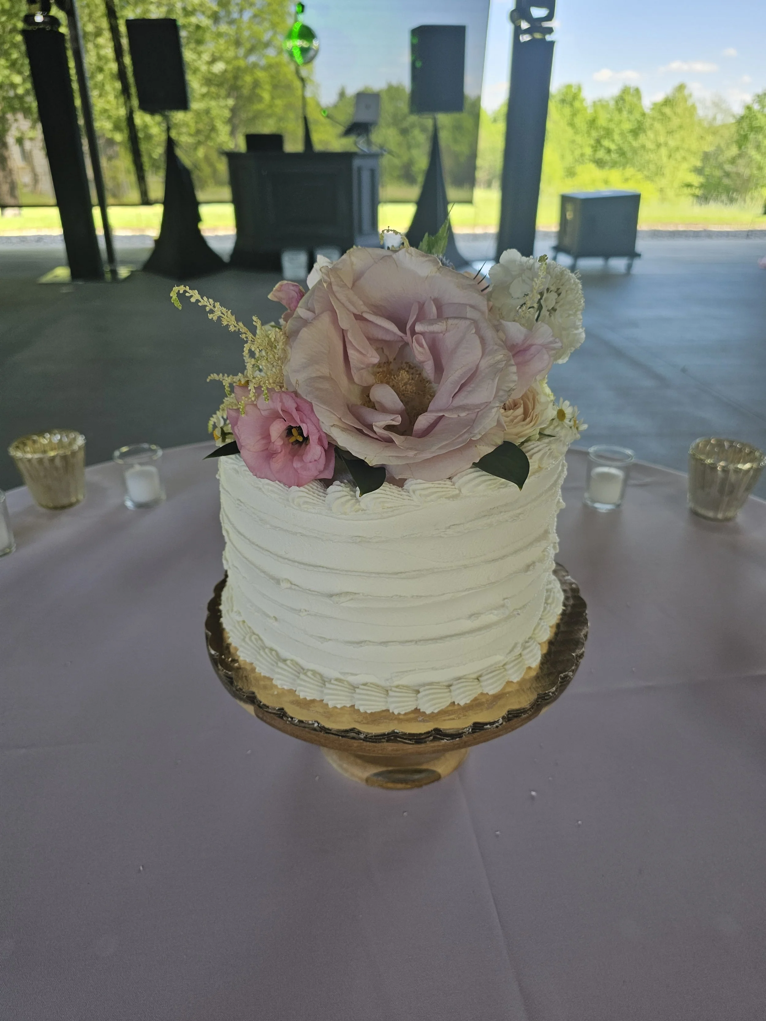 A white wedding cake with a pink and white floral topper on a table, simple knife edge design.