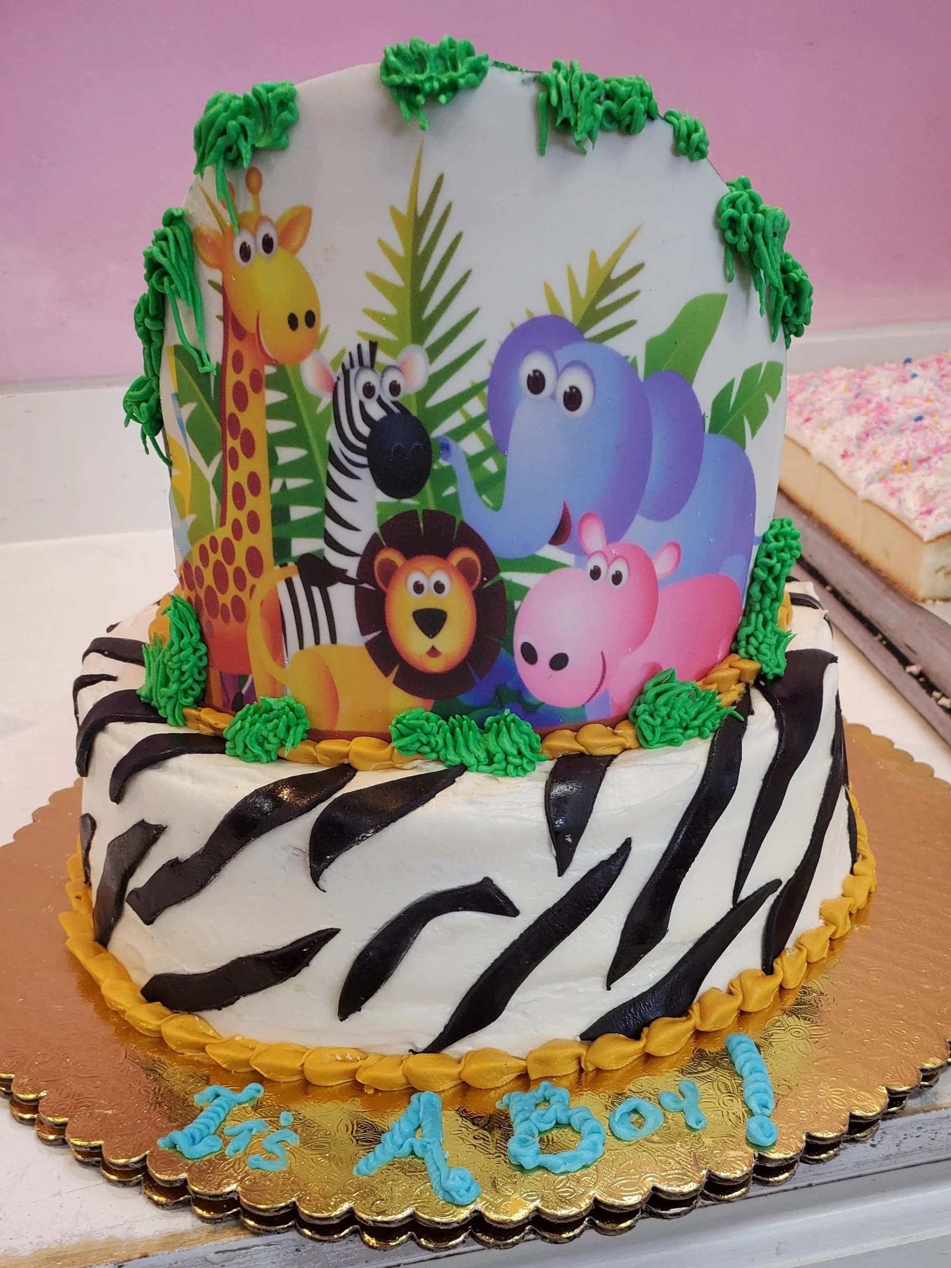 A two-tiered birthday cake with a zebra and jungle animal theme, featuring a printed topper of animals like a giraffe, zebra, lion, elephant, and hippopotamus with tropical leaves, decorated with black zebra print frosting and green, black, and yellow frosting accents.