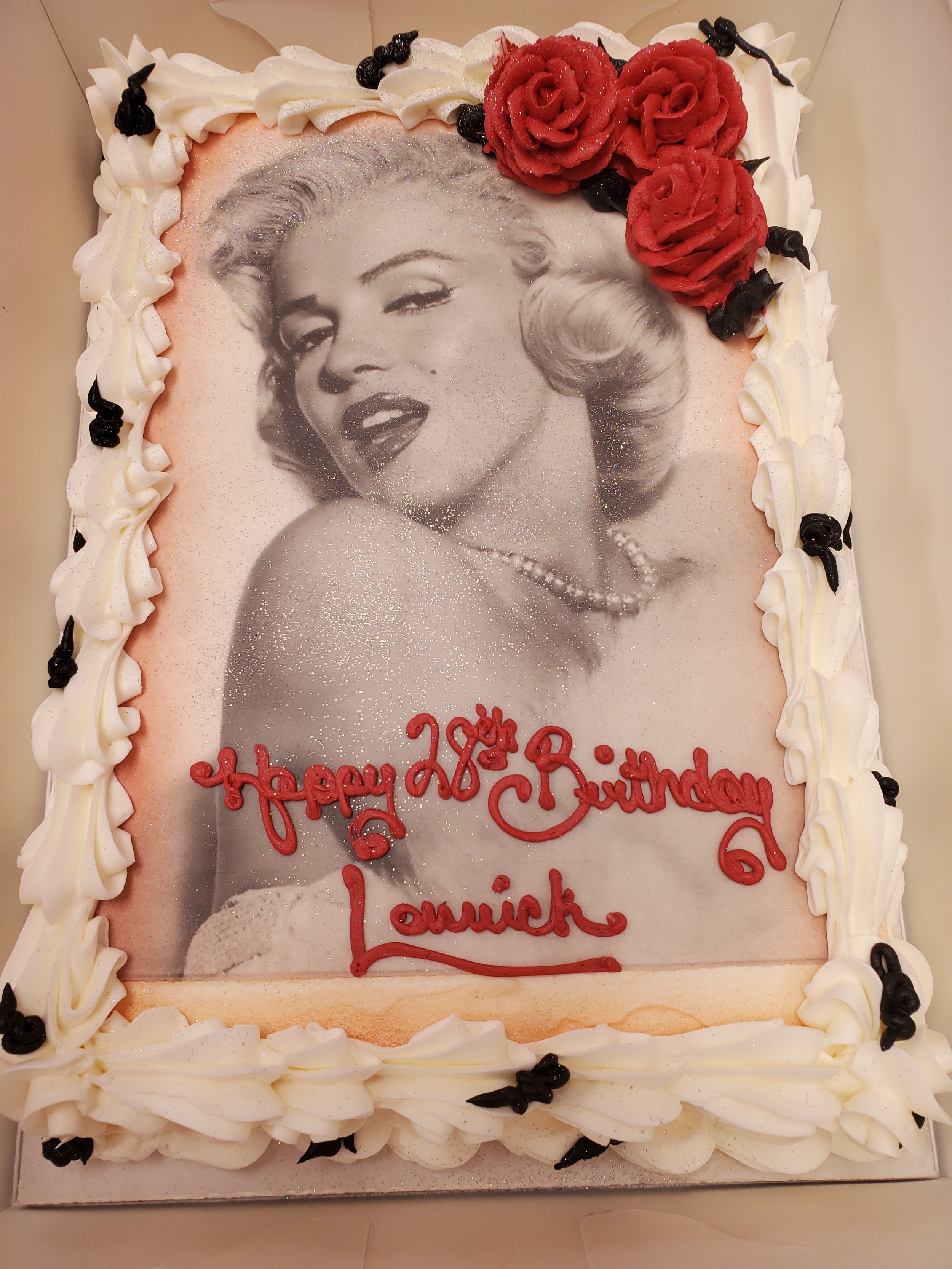Decorated birthday cake with a black and white photo of Marilyn Monroe printed on top, surrounded by white frosting, black accents, and red frosting roses, with red cursive writing that says "Happy 28th Birthday Louick."