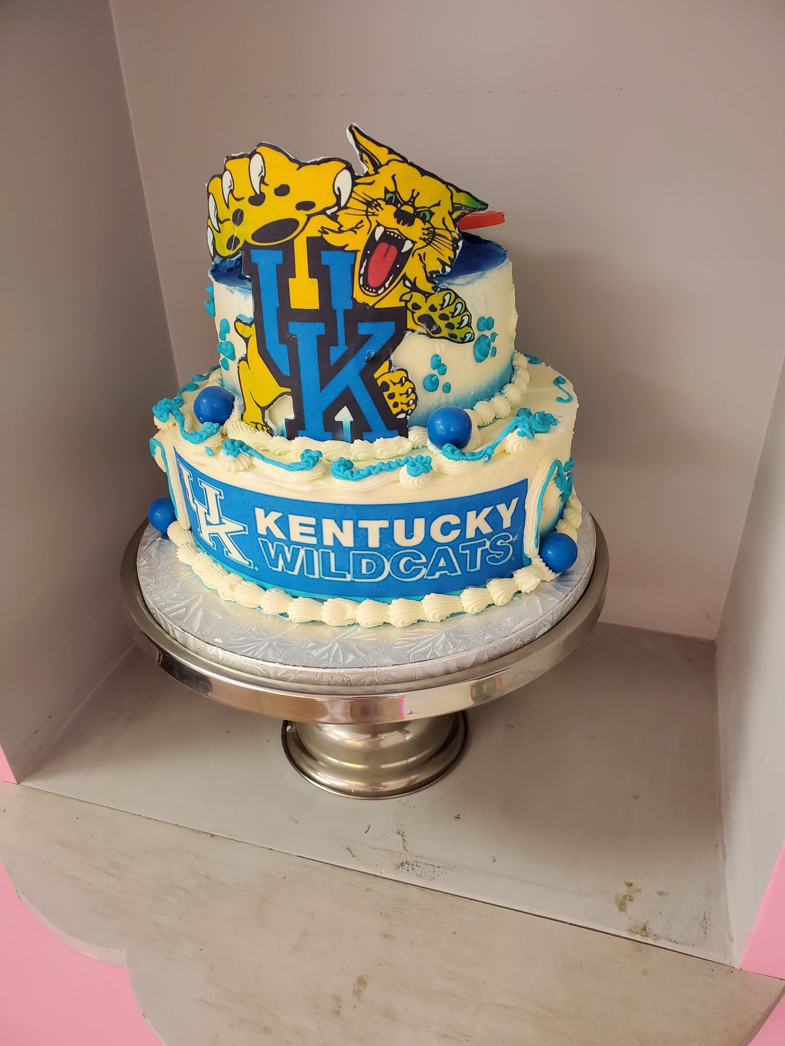 Cake with Kentucky Wildcats logo and mascot decorations, featuring blue and white frosting with blue balls and ribbon, displayed on a silver stand.