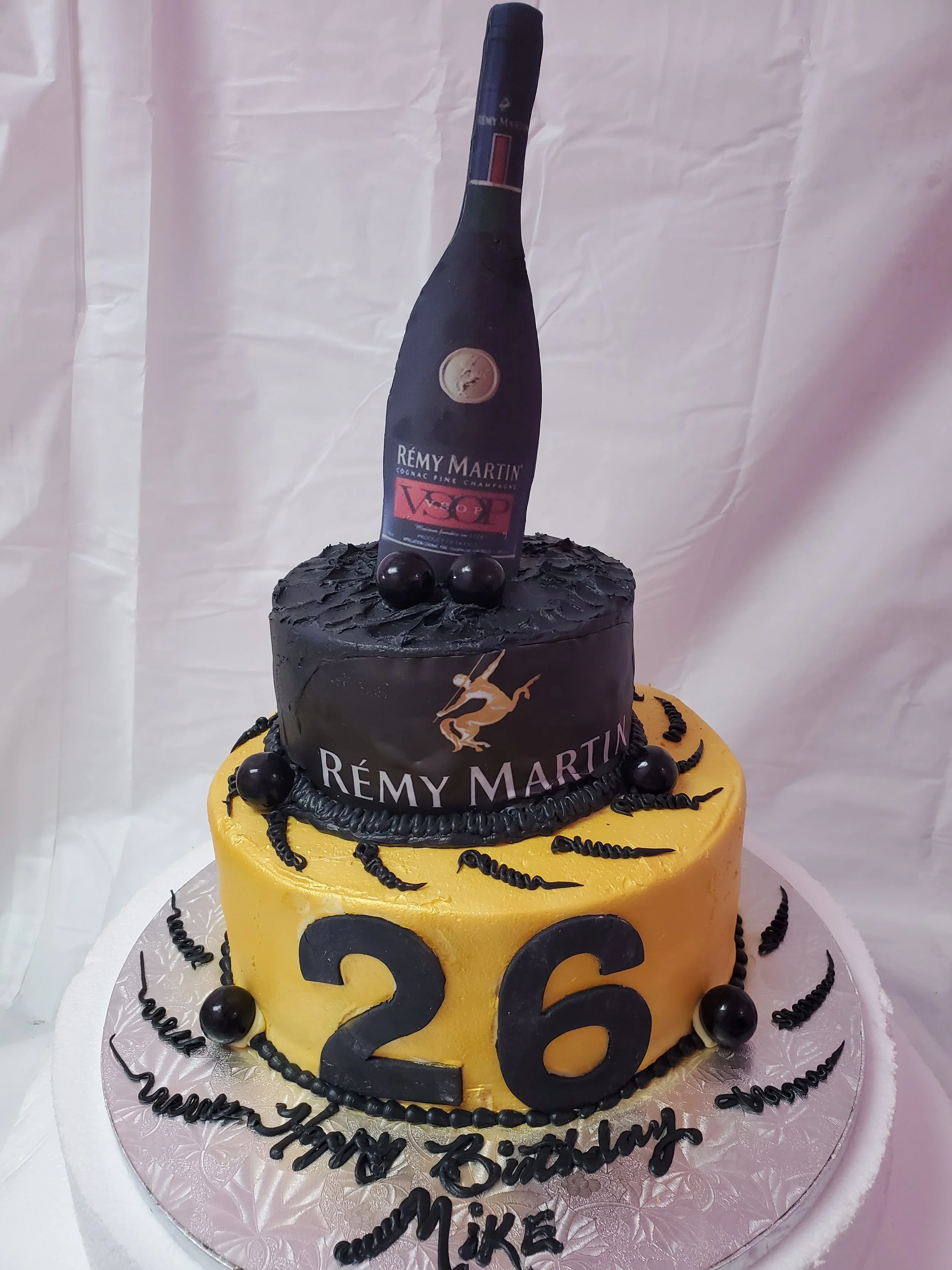 A two-tier birthday cake with gold and black decorations, featuring a bottle of Rémy Martin cognac with a red and black VIP label on top, and a large gold number 26 in front. The cake has black accents resembling worm-like shapes and black spheres around the edges, with a message written in black icing on the white base.