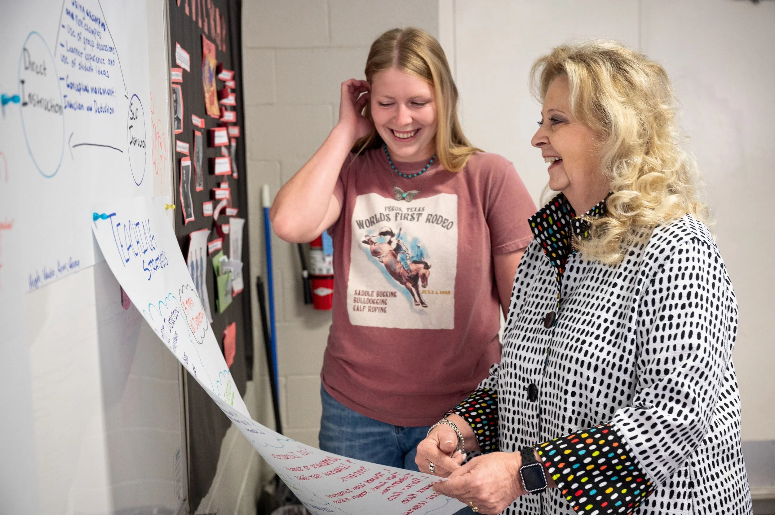 Two women, one young and one older, smiling and looking at a large poster with handwritten notes on a wall in a classroom or meeting room.