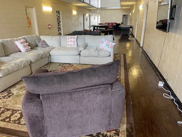 Living room with beige and white sofas, pink pillows, a patterned area rug, and dark hardwood floors, in front of a wall-mounted TV and with a hallway and dining area in the background.