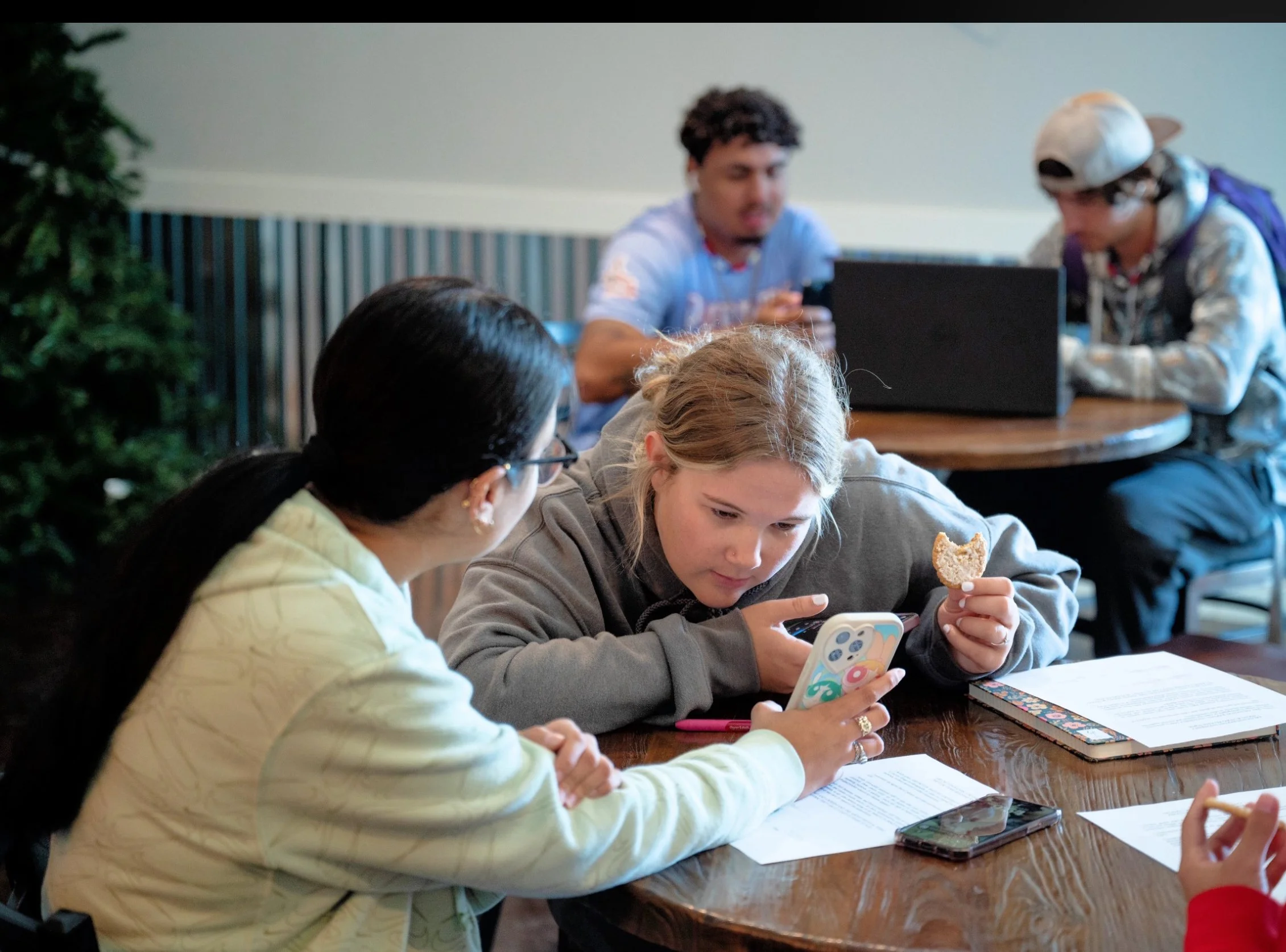 A group of young people sitting around a wooden table in a casual dining setting. One girl with blonde hair is looking at her phone, holding a cookie in her other hand. Another girl with dark hair and glasses is sitting beside her, looking at her. Th