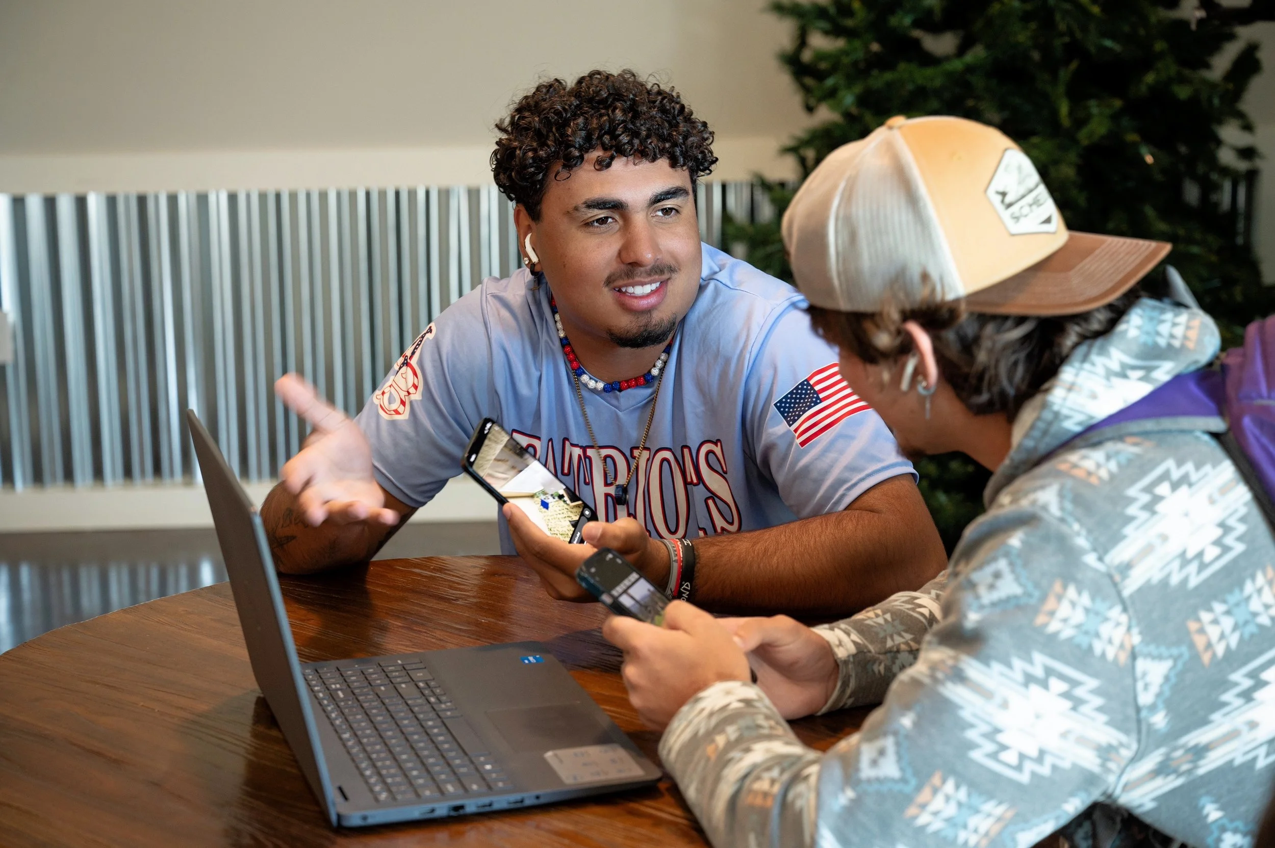 Two young men sitting at a wooden table, engaged in conversation, with laptops and smartphones in front of them. One is wearing a baseball cap and hoodie, the other is wearing a sports jersey and colorful beads, smiling and gesturing.