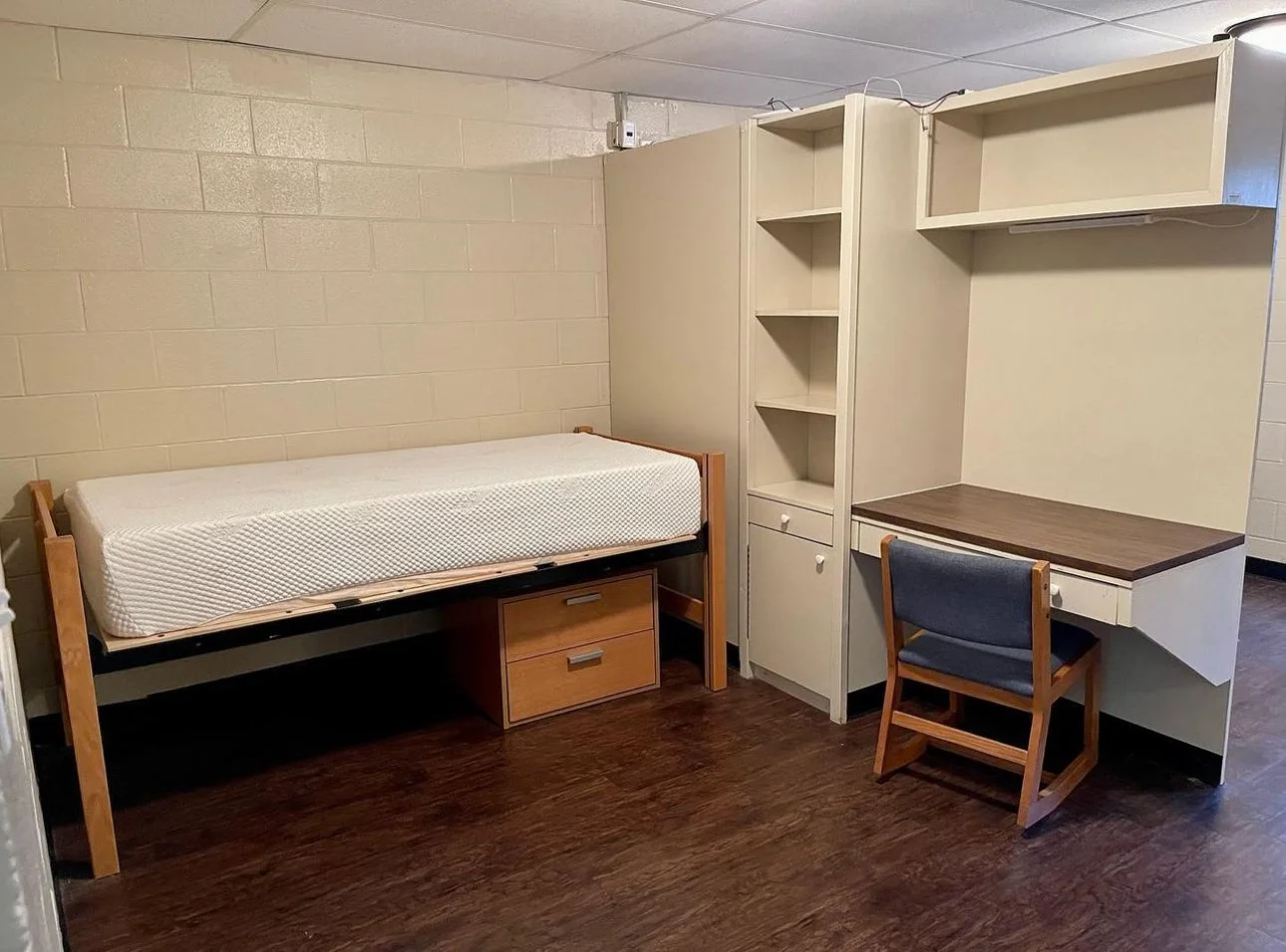 This furnished dorm room offers a simple, functional living space designed for comfort and study. Each resident has a twin bed with under-bed storage, a personal desk with shelving, and built-in closet space. The open layout provides ample room to mo