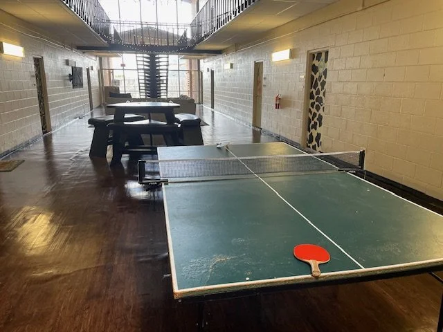 Indoor recreational room with a ping pong table in the foreground and a sitting area with tables and chairs at the back. Large windows at the far end allow natural light to illuminate the room.