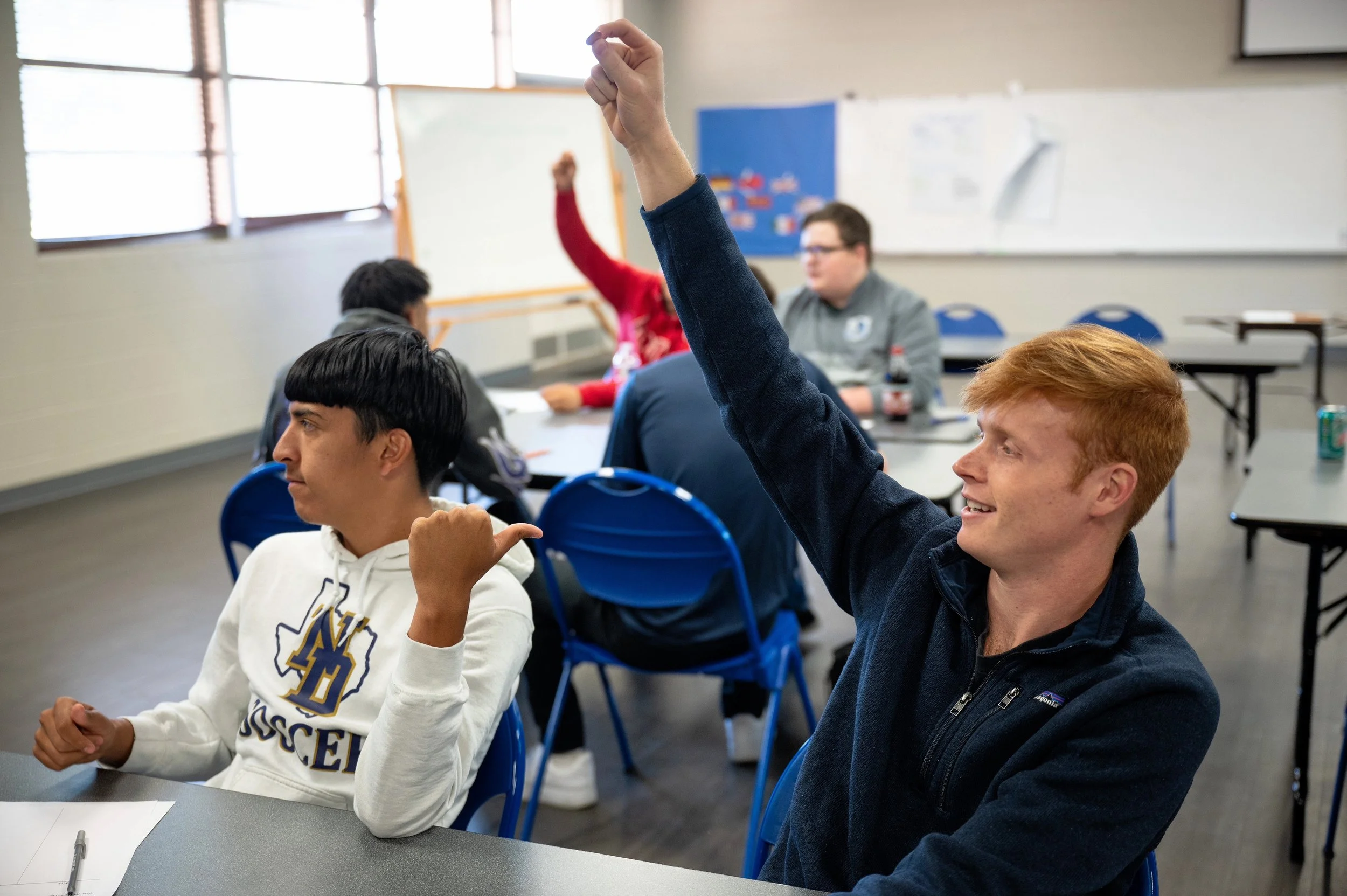 A classroom with students sitting at tables, one student with red hair raising their hand, and others listening, some with thumbs up gestures.