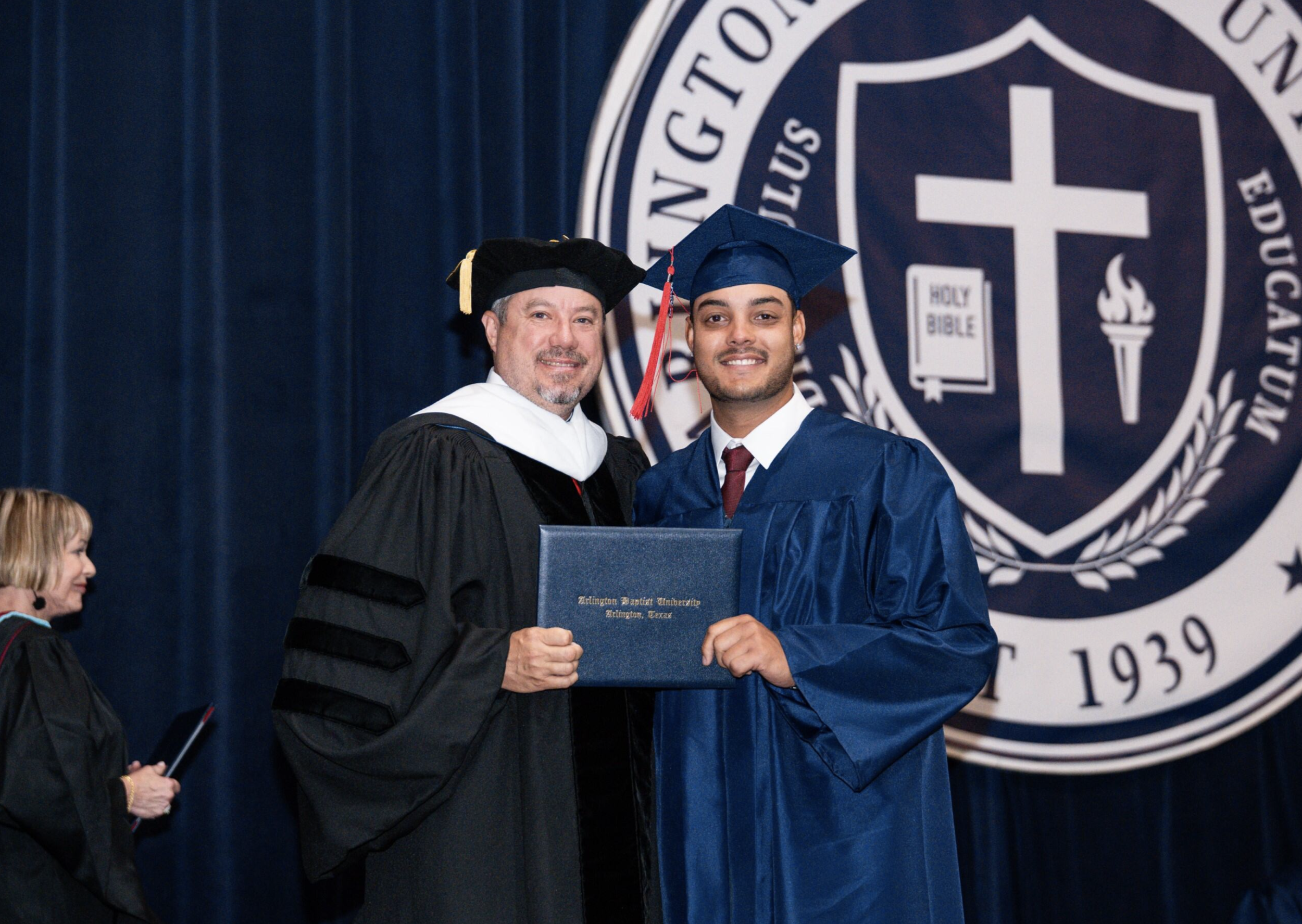 A male graduate in a navy blue cap and gown receiving a diploma from a university official in academic regalia on stage during graduation ceremony. The university emblem is visible in the background.