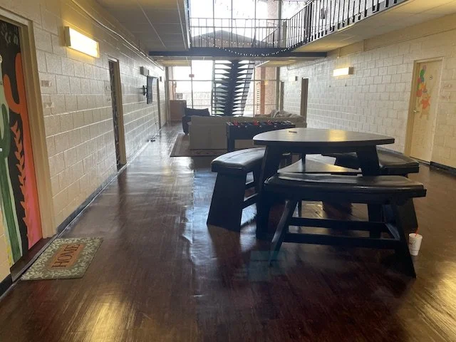 Indoor hallway with wooden flooring, featuring a high table with four benches in the foreground, a seating area with a foosball table and couches near the stairs, and a staircase leading to an upper level.