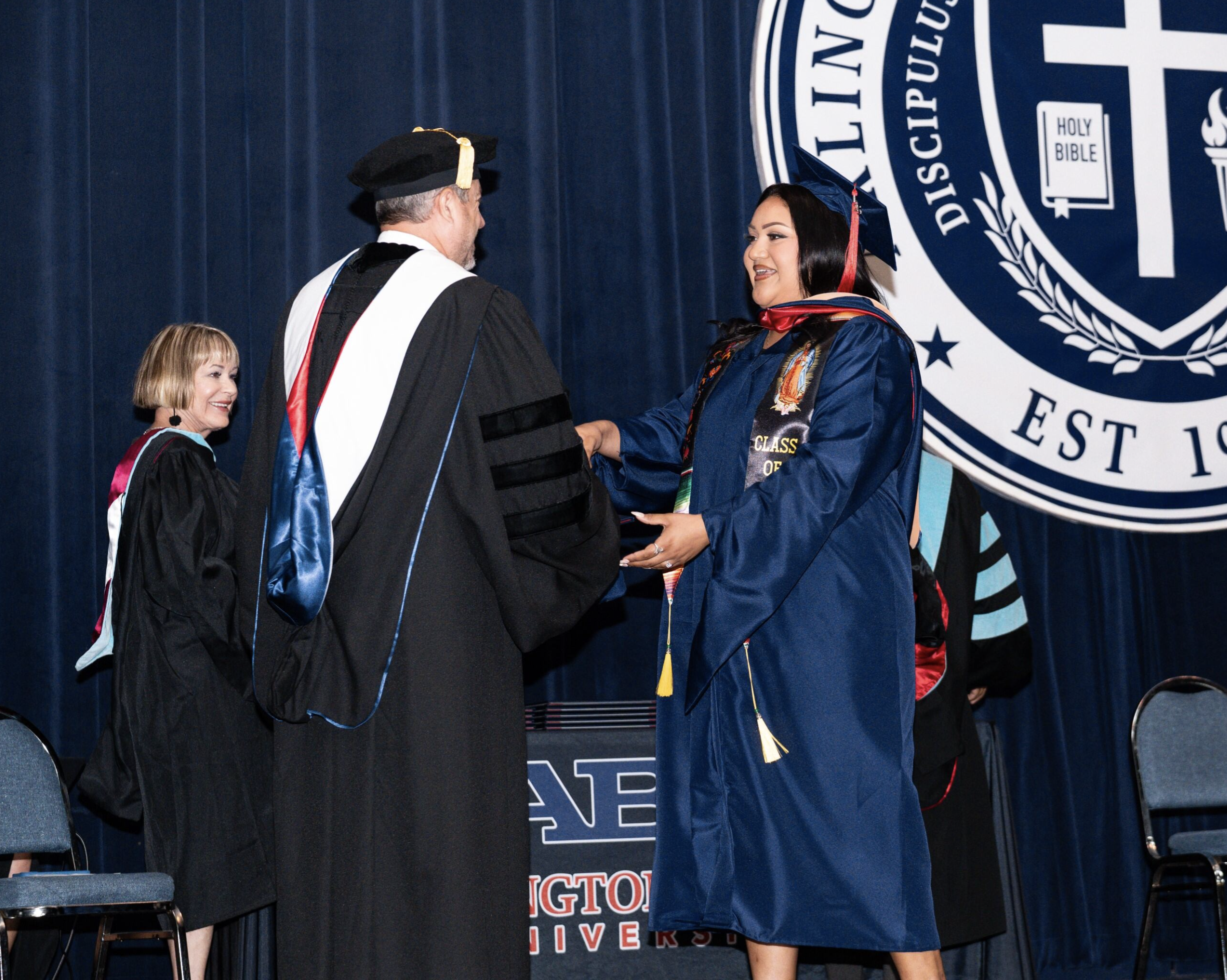 A graduate woman in blue cap and gown receiving diploma at college graduation ceremony, with others and official on stage, American university seal in background.