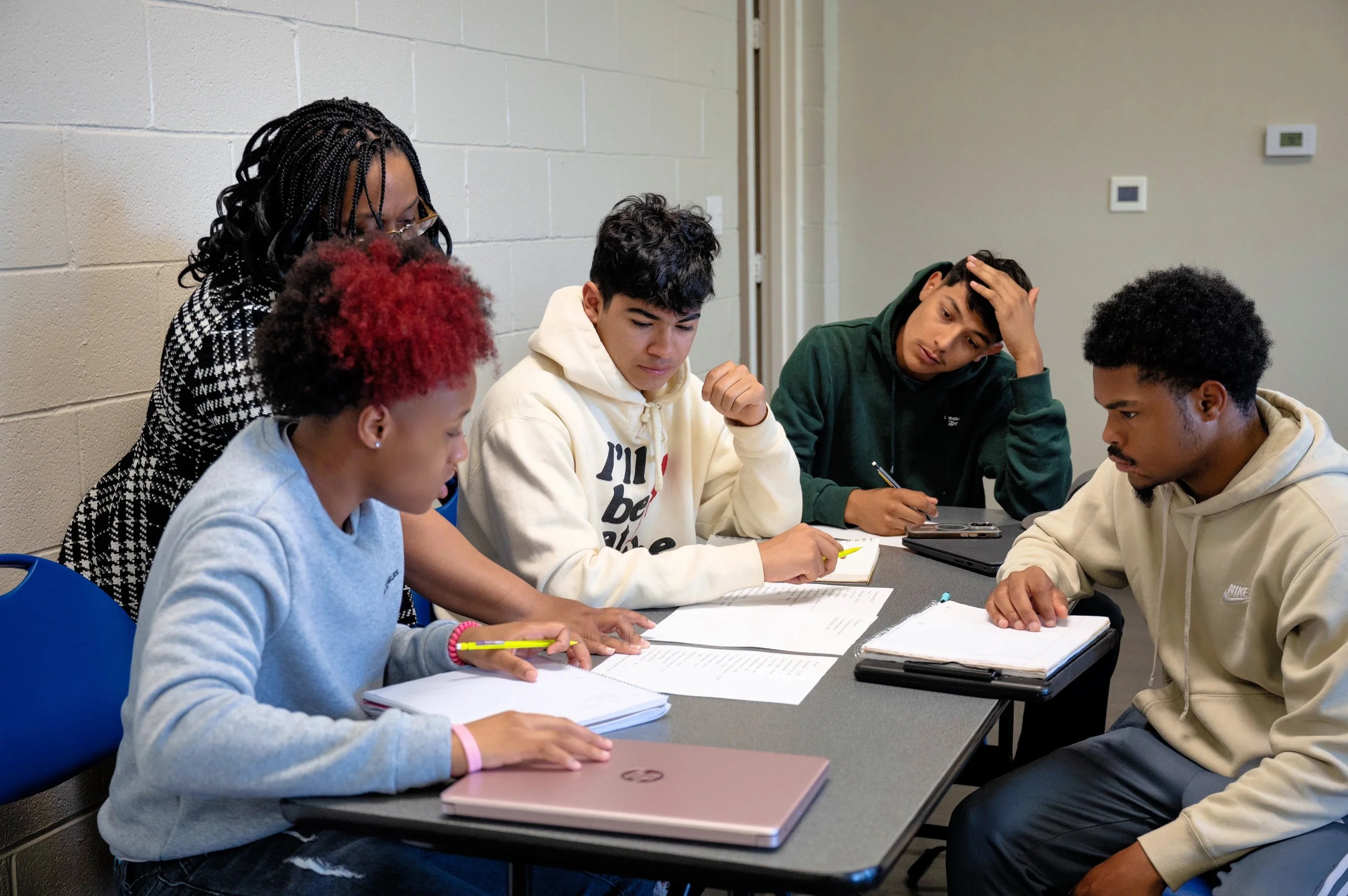 A group of five young people sitting at a table with notebooks and papers, working together in a classroom or study setting.