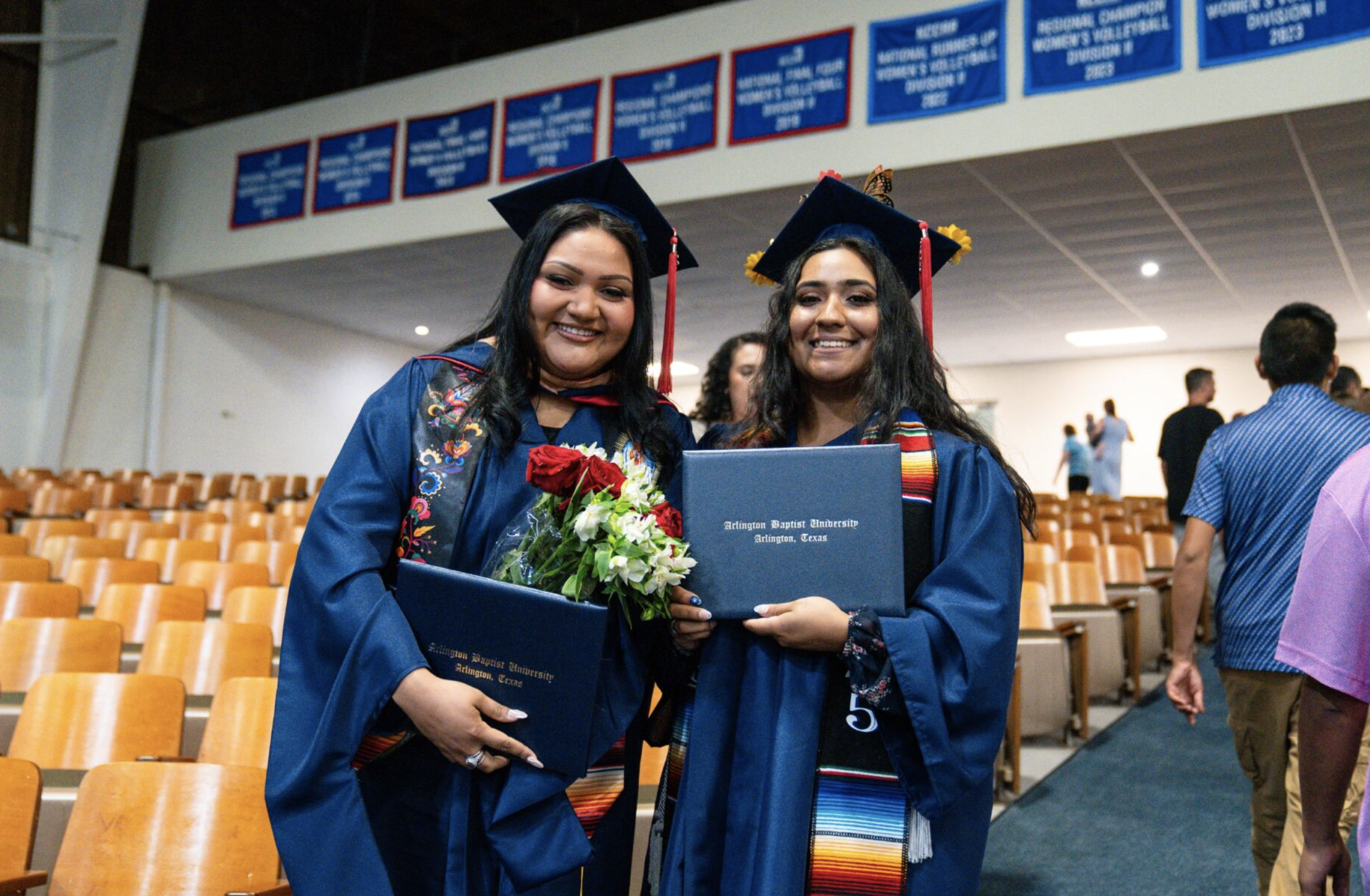 Two graduates in blue caps and gowns holding diplomas and flowers at a graduation ceremony inside an auditorium, with other attendees in the background.