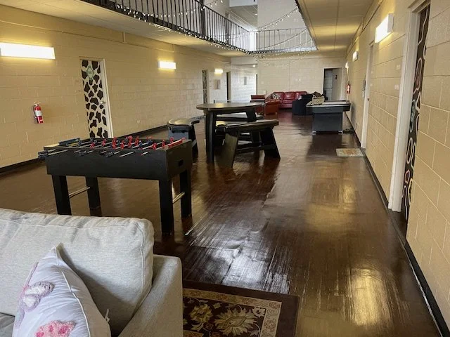 Interior of a recreational or lounge area with foosball, ping pong, and pool tables, and couch seating, with wooden flooring and beige walls.
