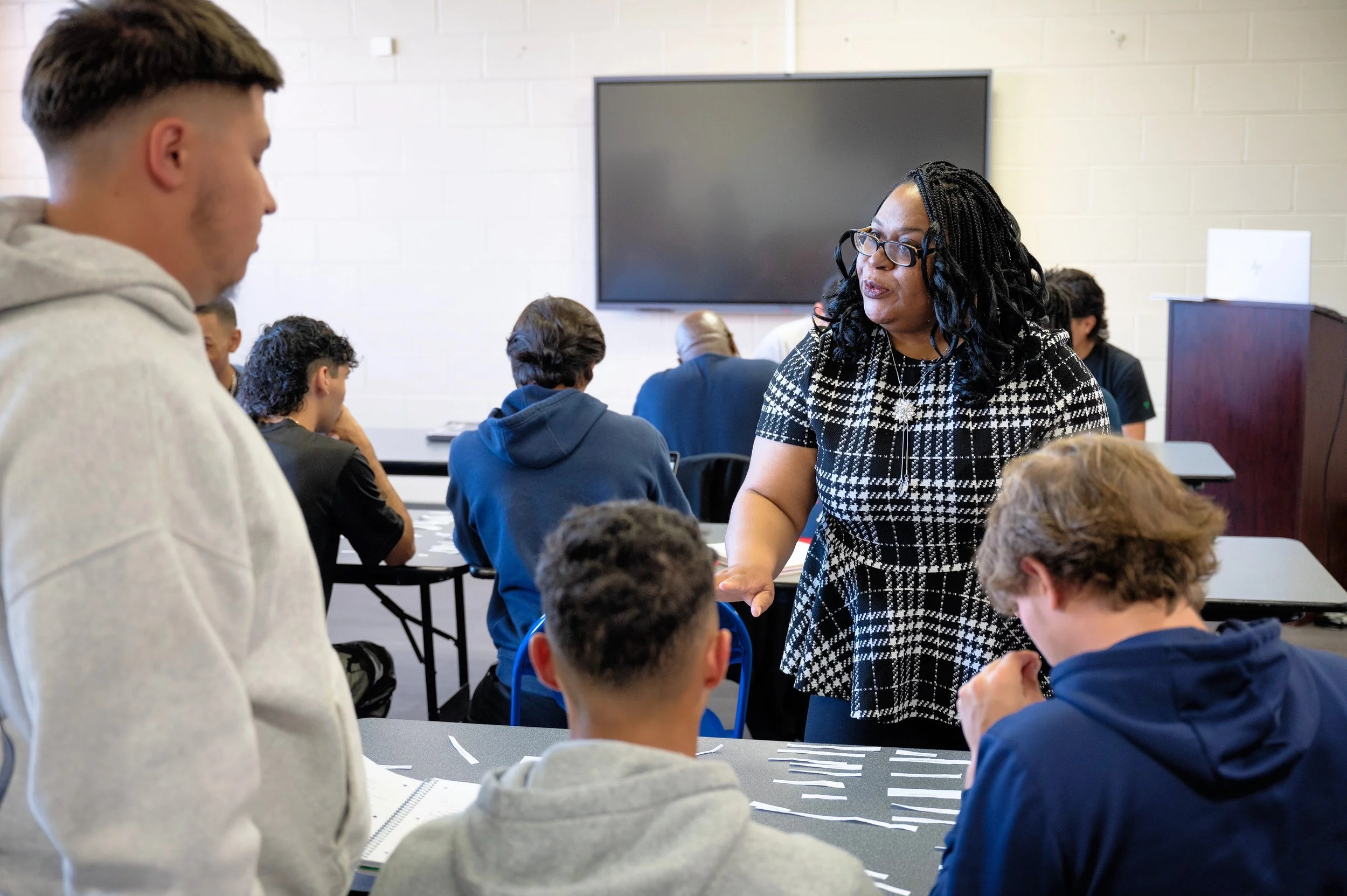 A classroom scene with a diverse group of students and a teacher. The teacher, a woman with long curly black hair and glasses, is speaking to students seated at tables. The students are engaged, some taking notes, with one standing and listening atte