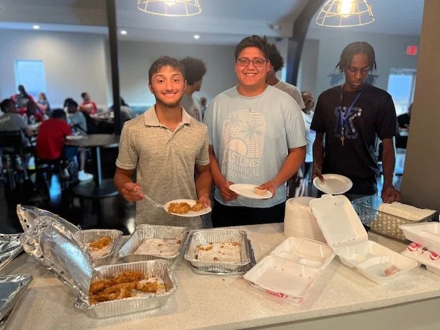 Three young men standing behind a buffet table with trays of fried chicken and plates of food, smiling at the camera in a busy dining hall.