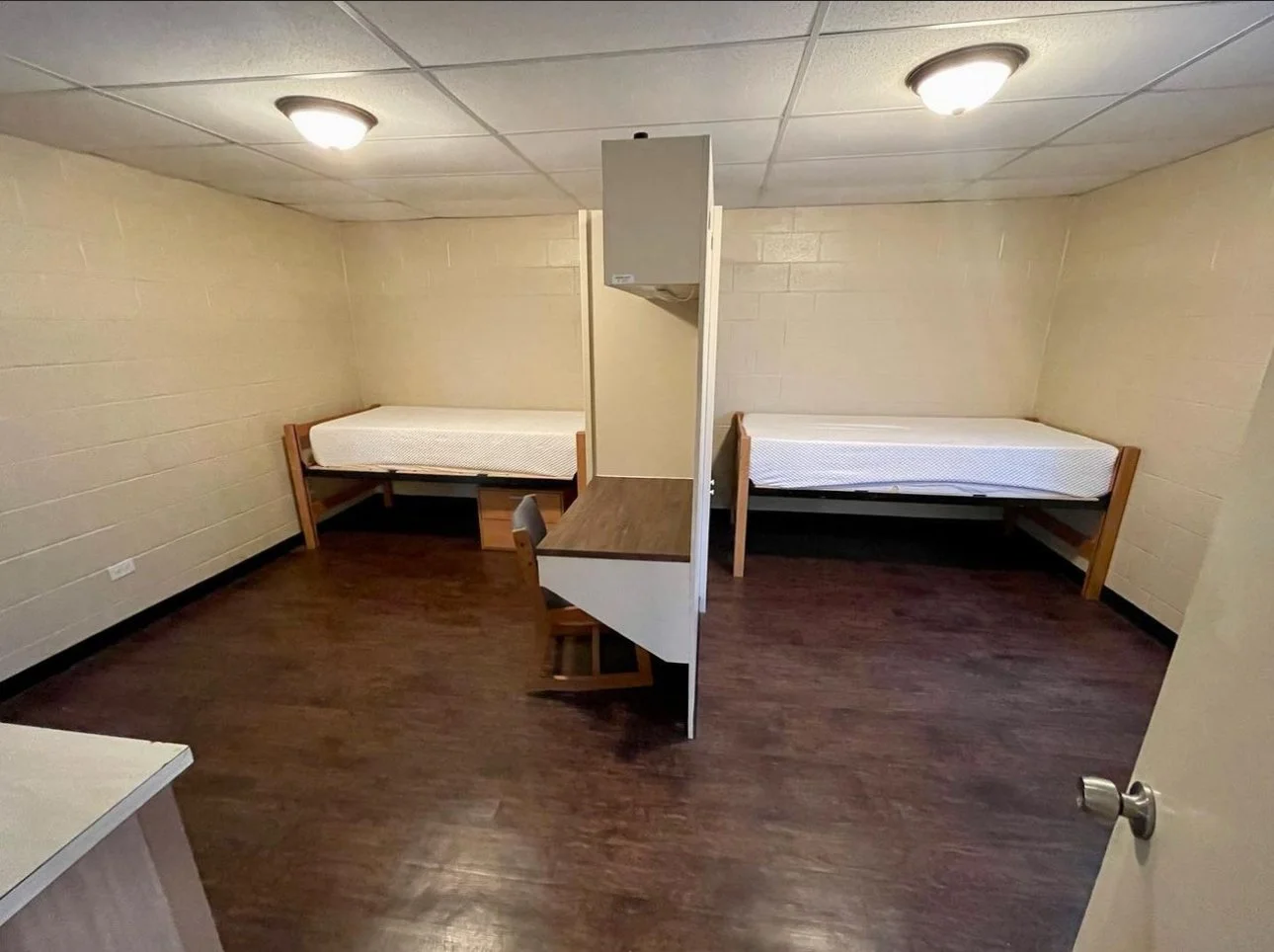 This furnished dorm room offers a simple, functional living space designed for comfort and study. Each resident has a twin bed with under-bed storage, a personal desk with shelving, and built-in closet space. The open layout provides ample room to mo