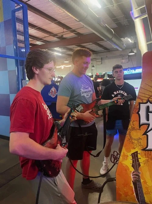 Three young men are playing a guitar-themed arcade game at an indoor entertainment center. The boy in red is holding a guitar controller, the boy in a gray T-shirt is also holding a guitar controller, and the boy in the background is watching.