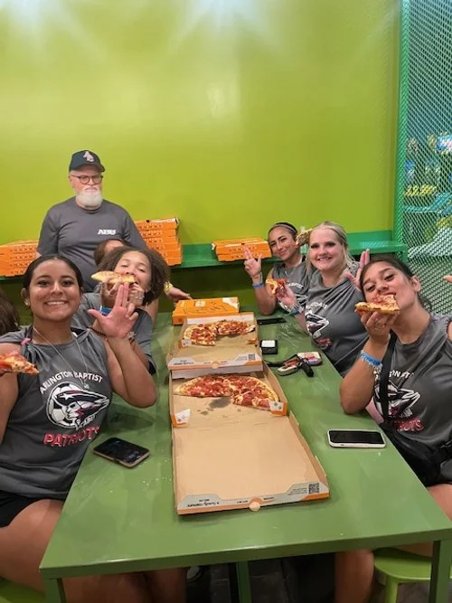 Group of six women and one man sitting at a green table inside a pizza eatery, holding slices of pizza and smiling. There are pizza boxes on the table, and the group appears to be celebrating after a meal.