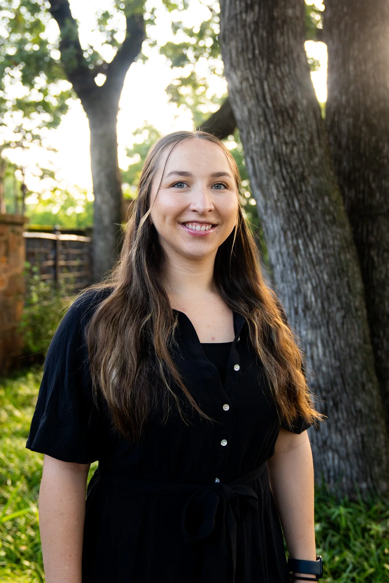 A young woman with long brown hair and blue eyes smiling outdoors near trees in a garden or park during daytime.