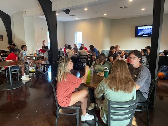 Children and teenagers sitting at tables in a room with a television on the wall.