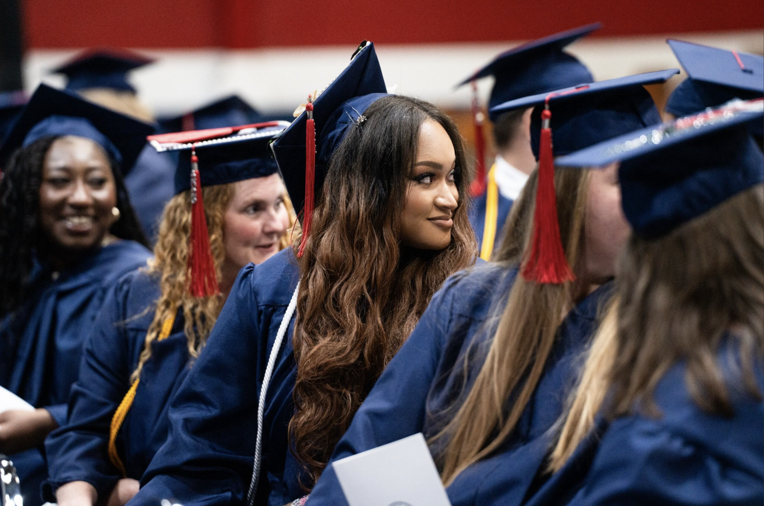 Group of graduates in blue caps and gowns sitting during a graduation ceremony, with one woman smiling and looking to the side.
