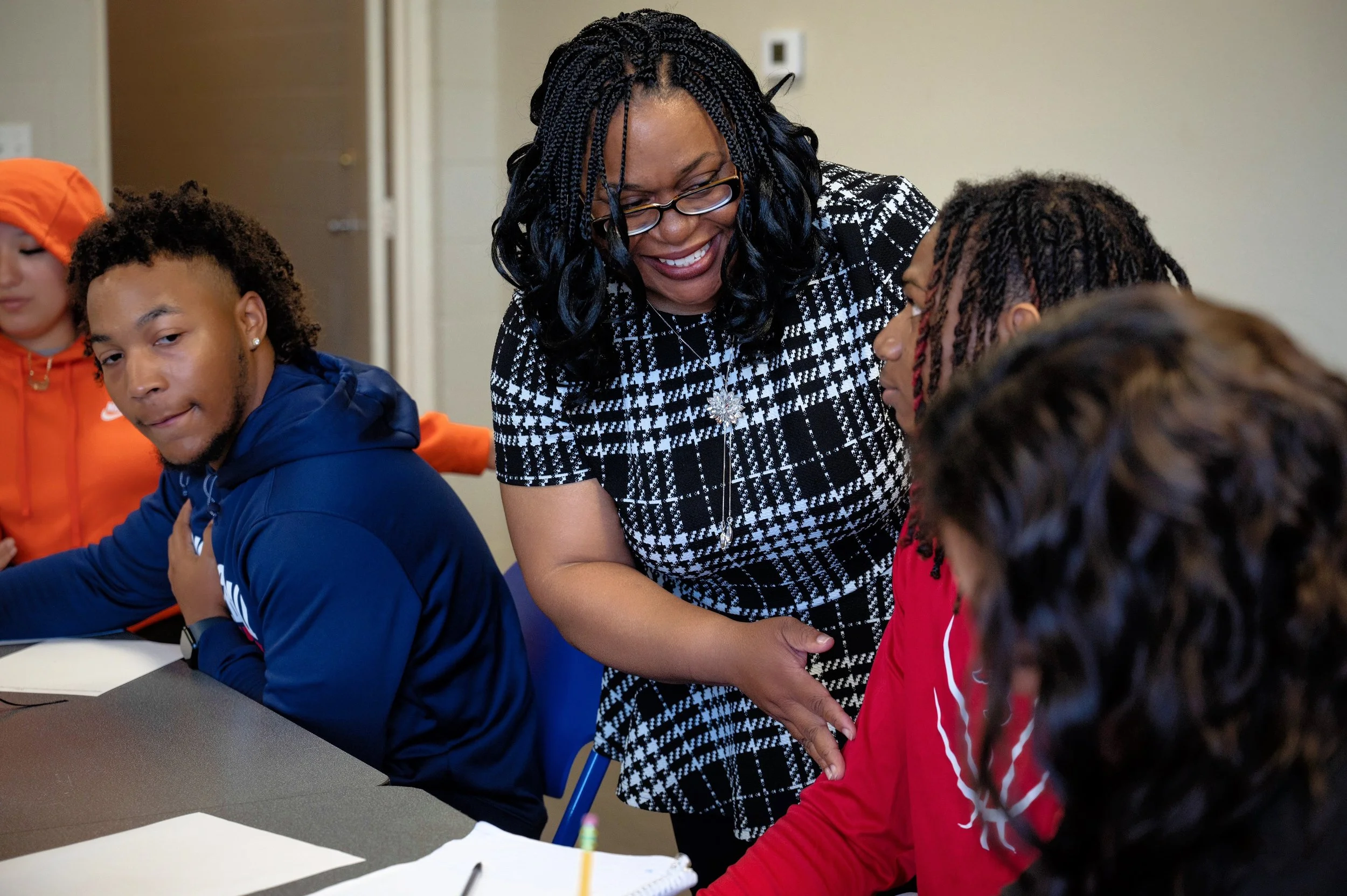 A smiling woman interacting with a group of young people seated at a table in a classroom or meeting room.