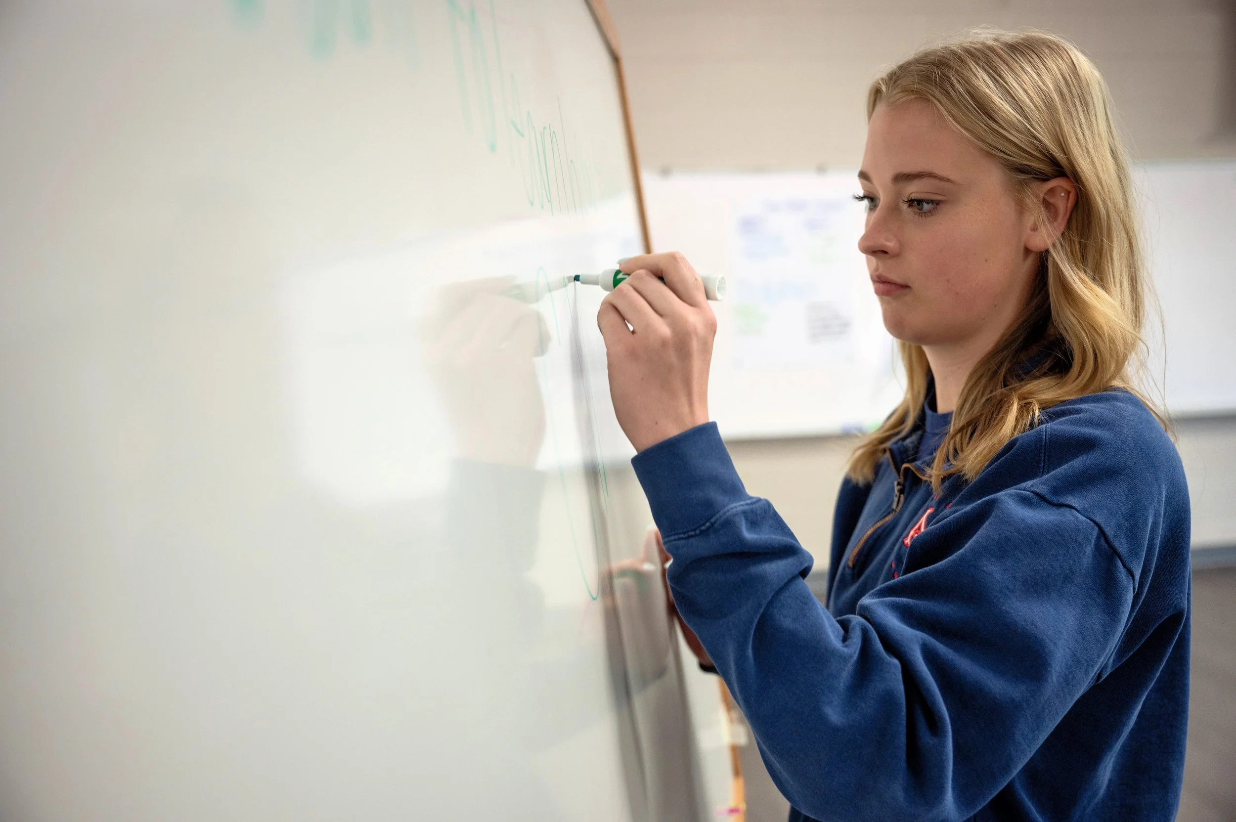 A young woman with blonde hair writes on a whiteboard with a green marker in a classroom or office setting.