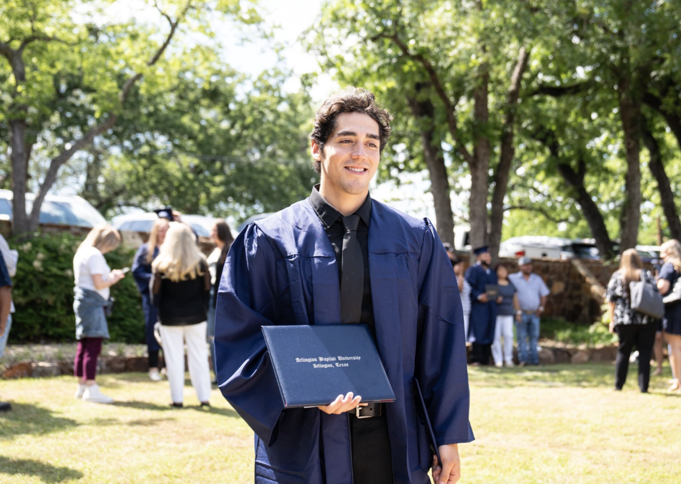 A young man in a blue graduation gown holding a diploma stands outdoors on a sunny day during graduation ceremony, with other graduates and spectators in the background.