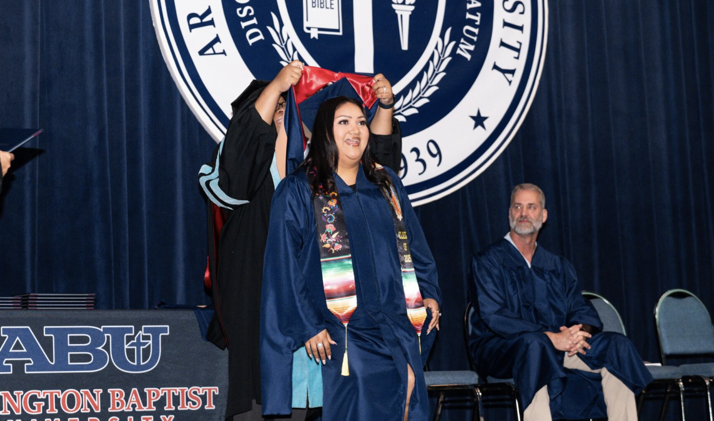 A graduation ceremony with a woman in a blue cap and gown standing on stage, being assisted into her cap by a staff member. A man in a blue gown is sitting to the right, observing. Behind them is a large emblem of a Baptist church on a dark blue curt