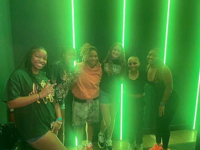 Group of six smiling young women posing together in front of green neon lights.