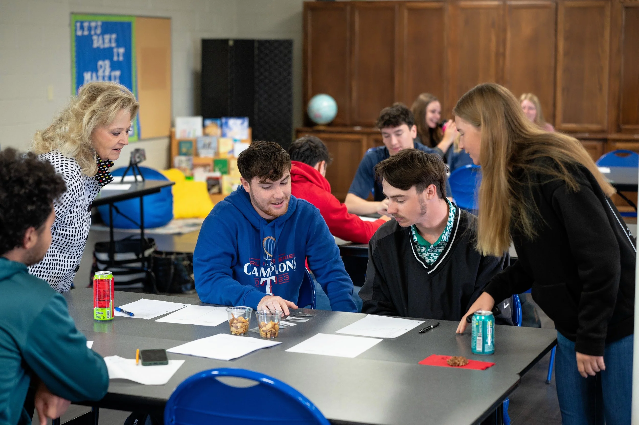 Teacher guiding high school students in a classroom during a group activity with snacks and papers on table.