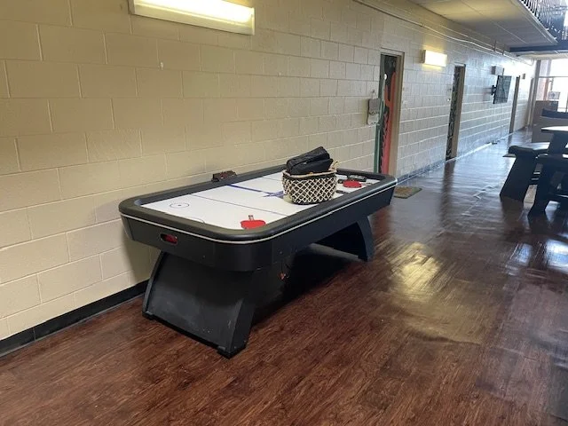Indoor hallway with a small boat-shaped air hockey table against a beige brick wall, basket on top, and folded black cloth inside.