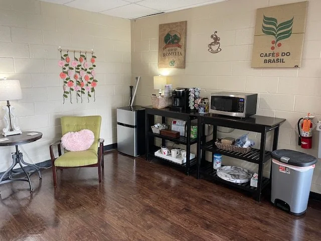 A small coffee station with a microwave, mini fridge, and shelves holding cups, plates, and supplies. There is a green armchair with a pink fluffy pillow, a side table with a lamp, and wall art decorated with coffee-themed designs.