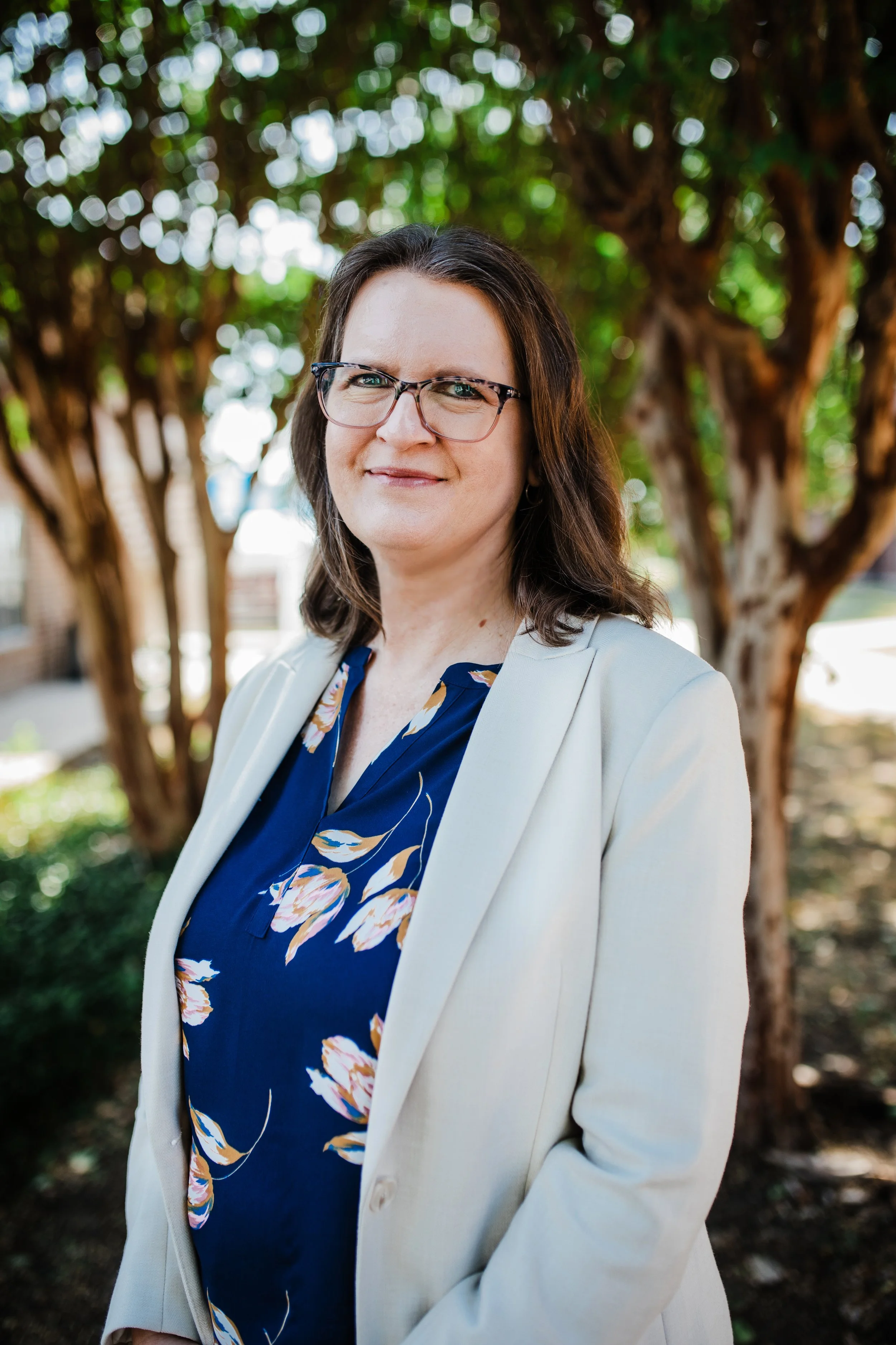 A woman with shoulder-length brown hair, glasses, wearing a black blazer and a red blouse with white patterns, standing against a textured blue background.