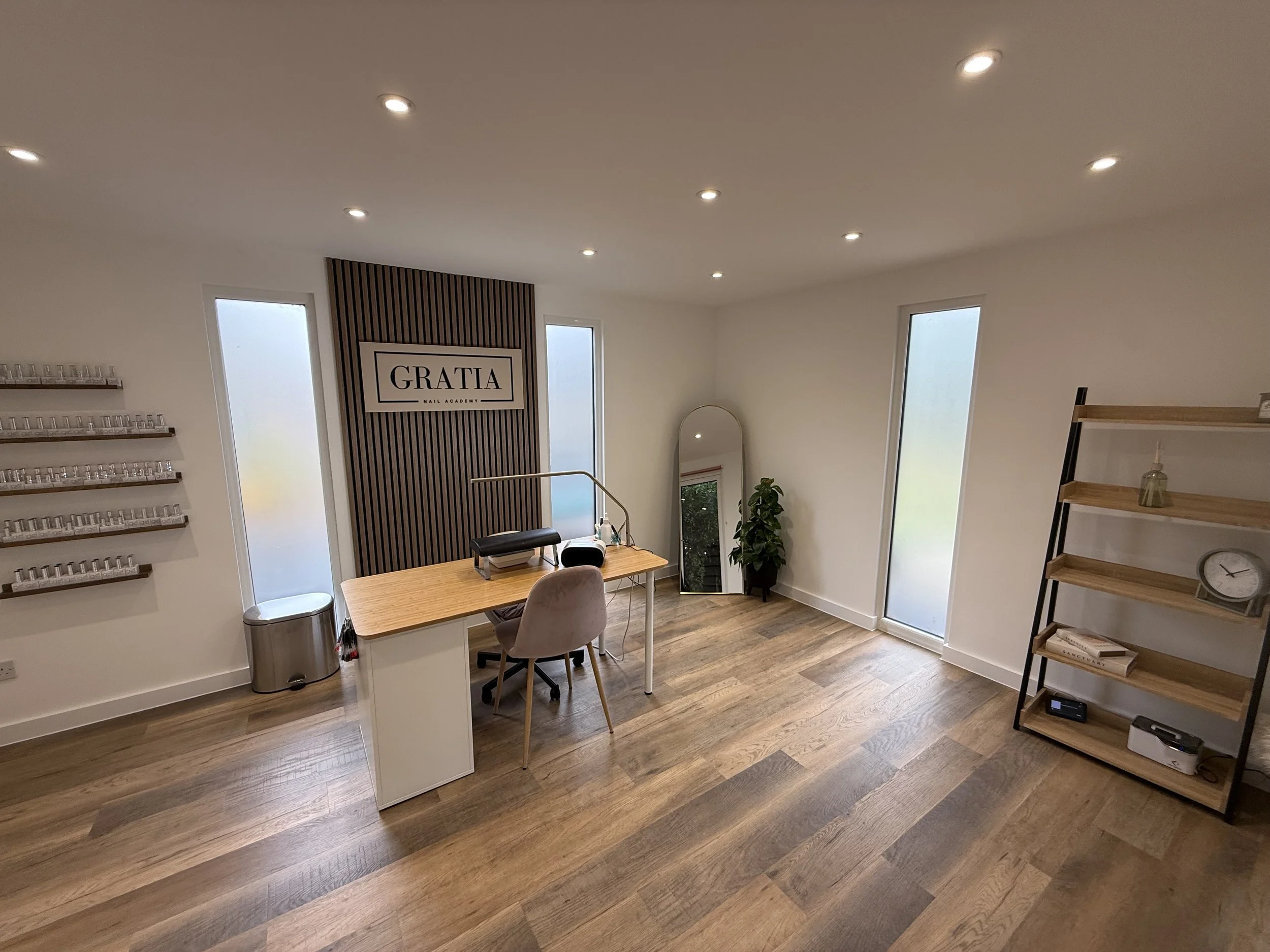 Interior of a nail salon with a wooden floor, a white reception desk, a wall with framed nail polish samples, a standing mirror, and shelves with decor and books. Sign reads 'Gratia Nail Academy'.