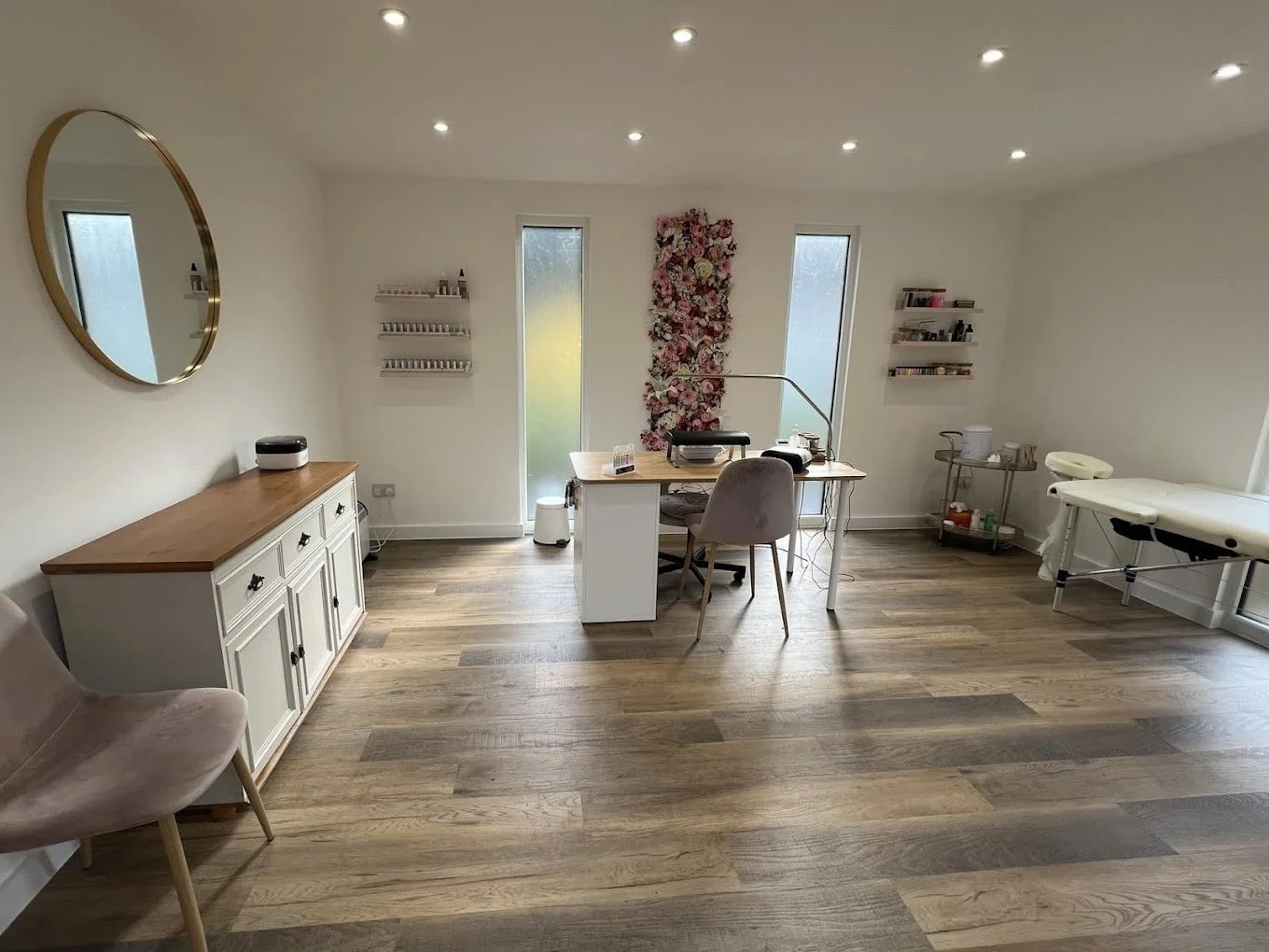 A spacious, well-lit treatment room with wooden floors, featuring a massage table, a desk with a chair, shelves with beauty products, a round mirror on the wall, and a floral wall decoration.