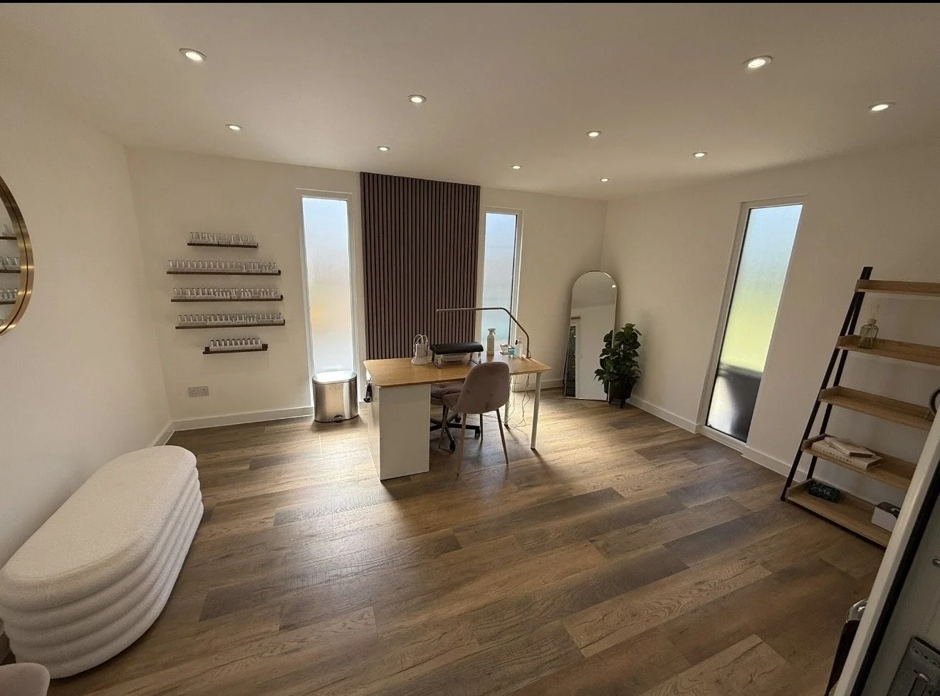 A modern, minimally decorated room with wood flooring, a desk with a chair, a large mirror, a shelving unit with books, and tall windows allowing natural light.
