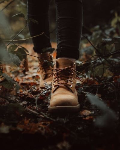 Close-up of a person's tan leather boots walking through fallen leaves and twigs in a forest or wooded area.