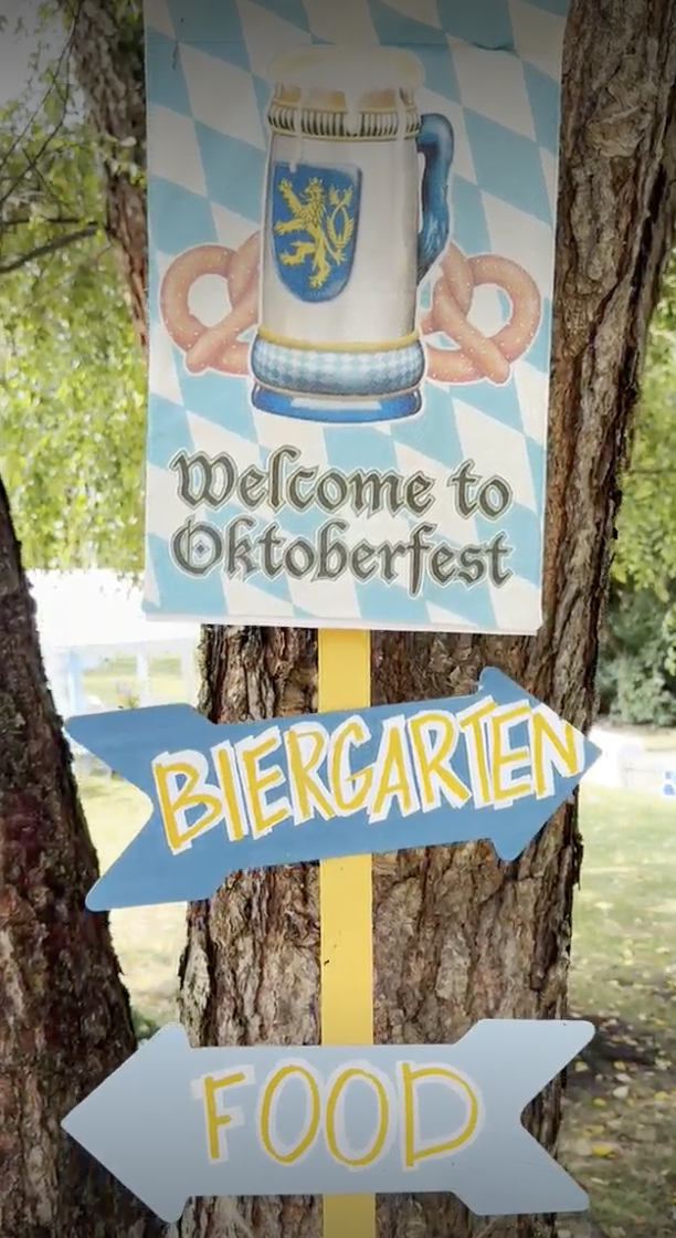 Signs attached to a tree, one saying "Welcome to Oktoberfest" with an illustration of a beer stein, and two directional signs labeled "Biergarten" pointing right and "Food" pointing left, in an outdoor setting.
