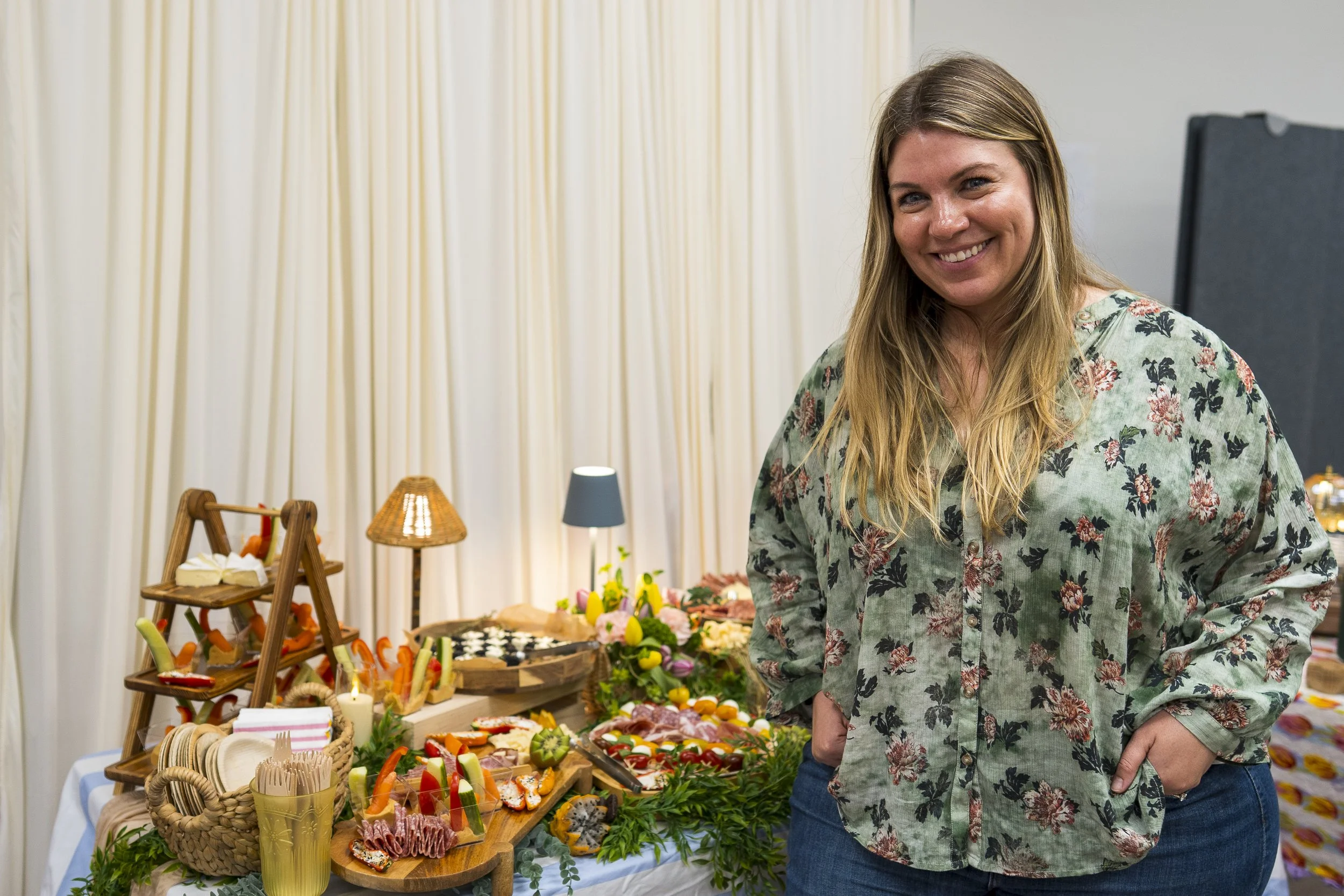 A woman with long blonde hair, wearing a green floral shirt, smiling at a table with various colorful appetizers and snacks, and a display of decorative food items behind her in a well-lit room.