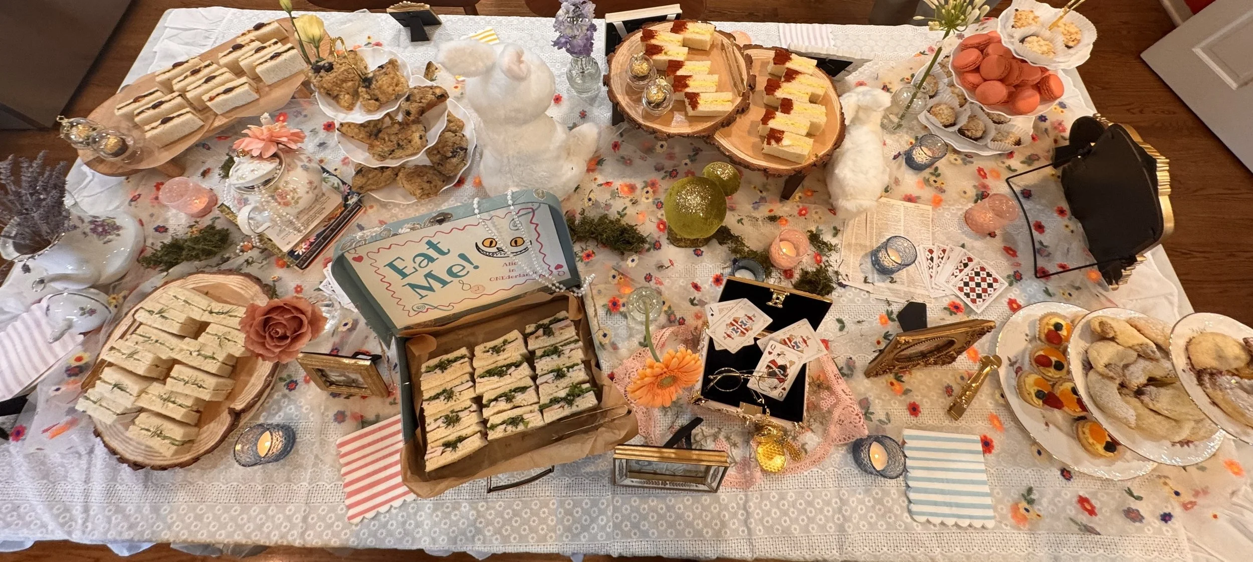 A table decorated with a floral tablecloth and various desserts, sandwiches, and decorations for a celebration, including cupcakes, macarons, cookies, sandwich trays, flowers, candles, playing cards, and a sign that says 'Eat Me!'