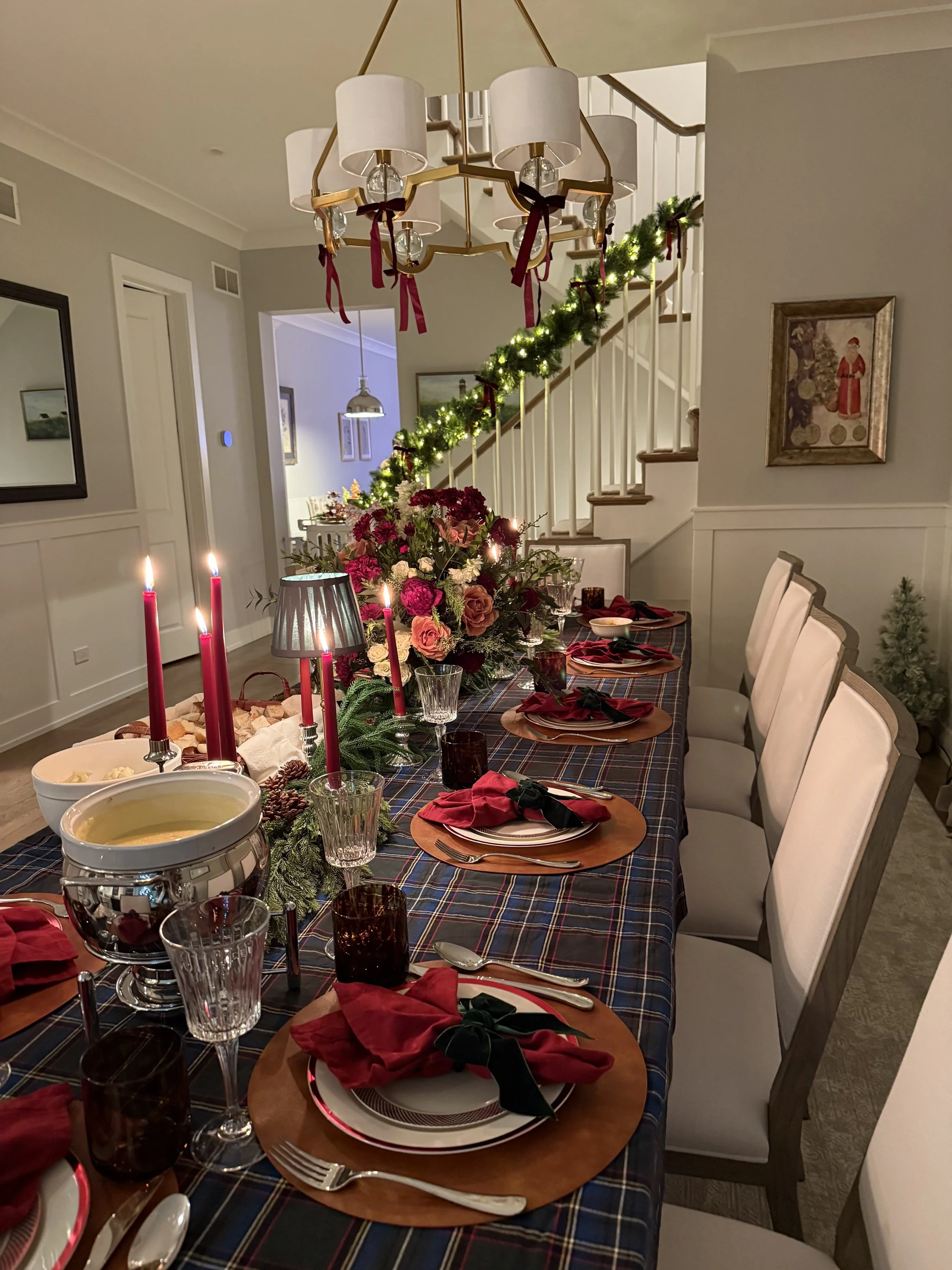 A dining room decorated for Christmas with a long table covered in a dark plaid tablecloth, set with plates, wine glasses, and red napkins tied with black ribbons. A large floral centerpiece with red and white flowers and candles is in the middle. A green garland with lights and bows hangs along the staircase railing. A chandelier with white shades hangs from the ceiling. The room has white walls with framed artwork and a mirror, and warm lighting creates a cozy holiday atmosphere.